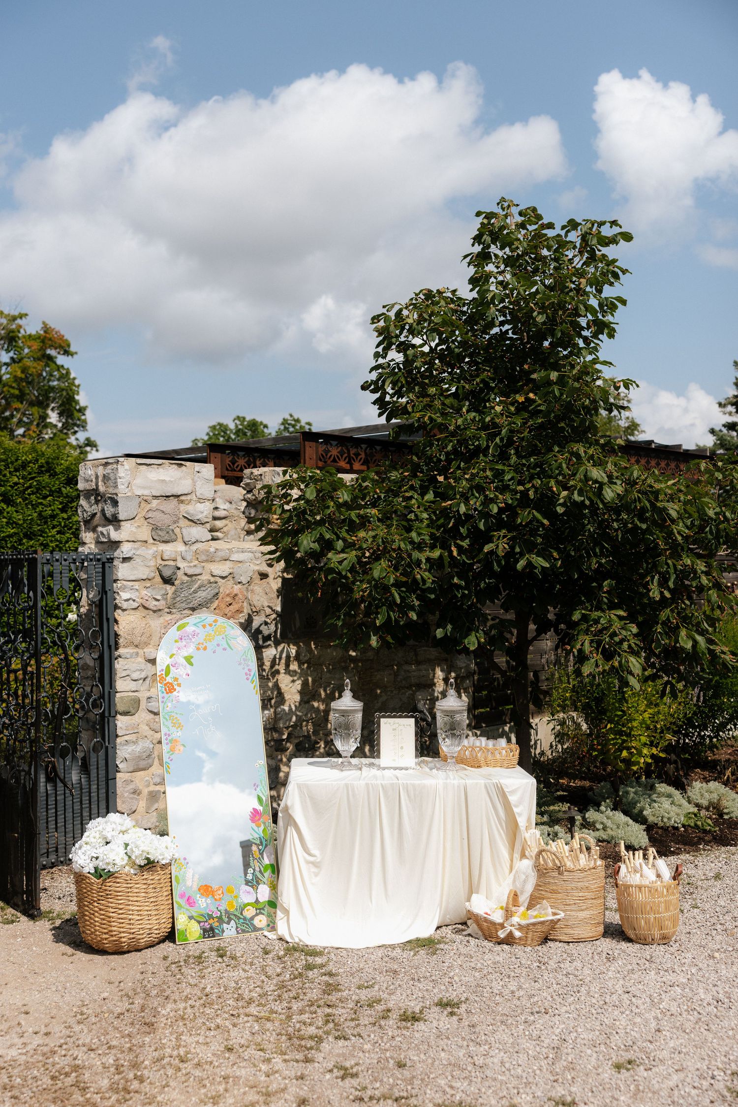 Outdoor wedding welcome table with white linen, wicker baskets, floral sign, and lanterns set against a stone wall.