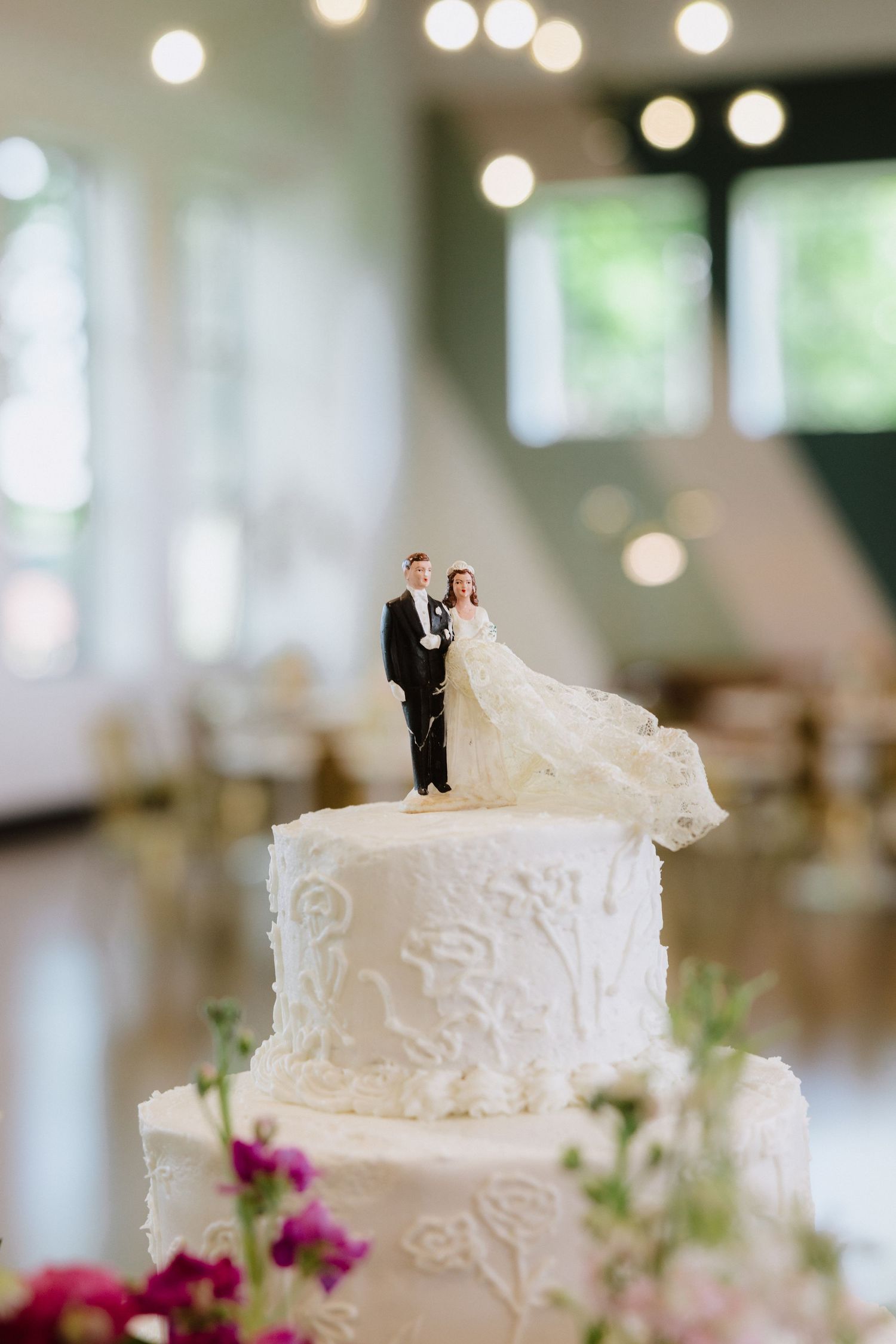 White tiered wedding cake with classic bride and groom figurine topper, decorated with floral lace details in elegant reception hall.