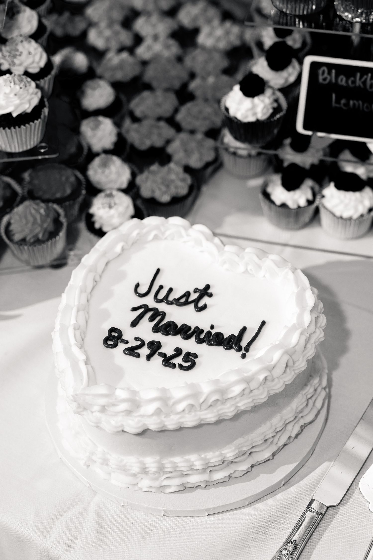 Heart-shaped Just Married wedding cake dated 8-29-25 with white frosting, displayed beside assorted cupcakes on a dessert table.