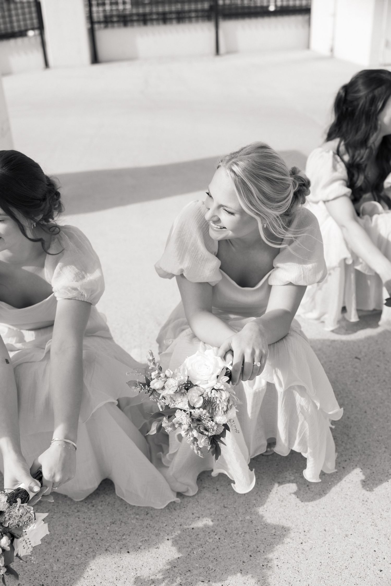 Black and white photo of bridesmaids in white dresses sitting together, smiling and holding bouquets.
