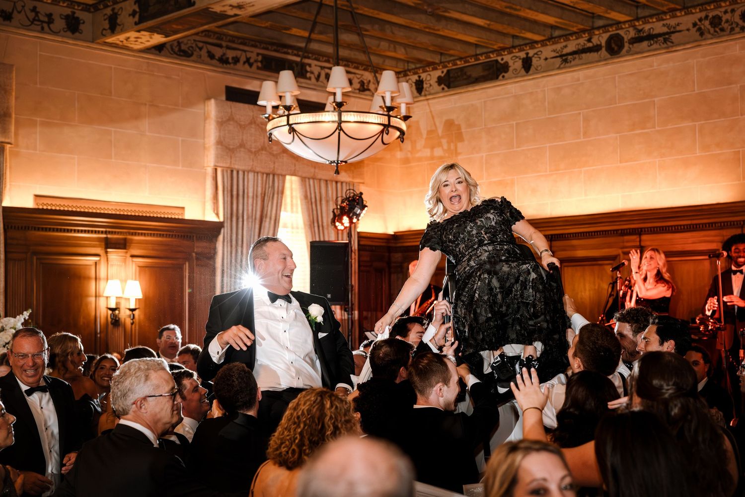 Woman in black dress lifted by crowd during energetic hora dance at wedding.