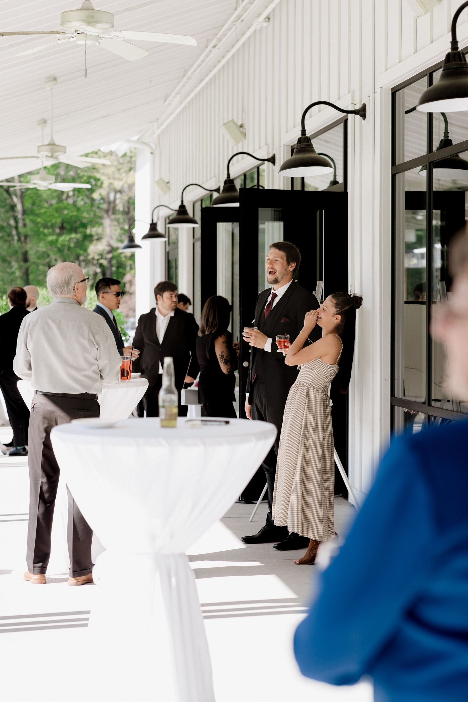 Wedding guests mingle on a bright white covered porch with cocktail tables and black barn lights during a reception.