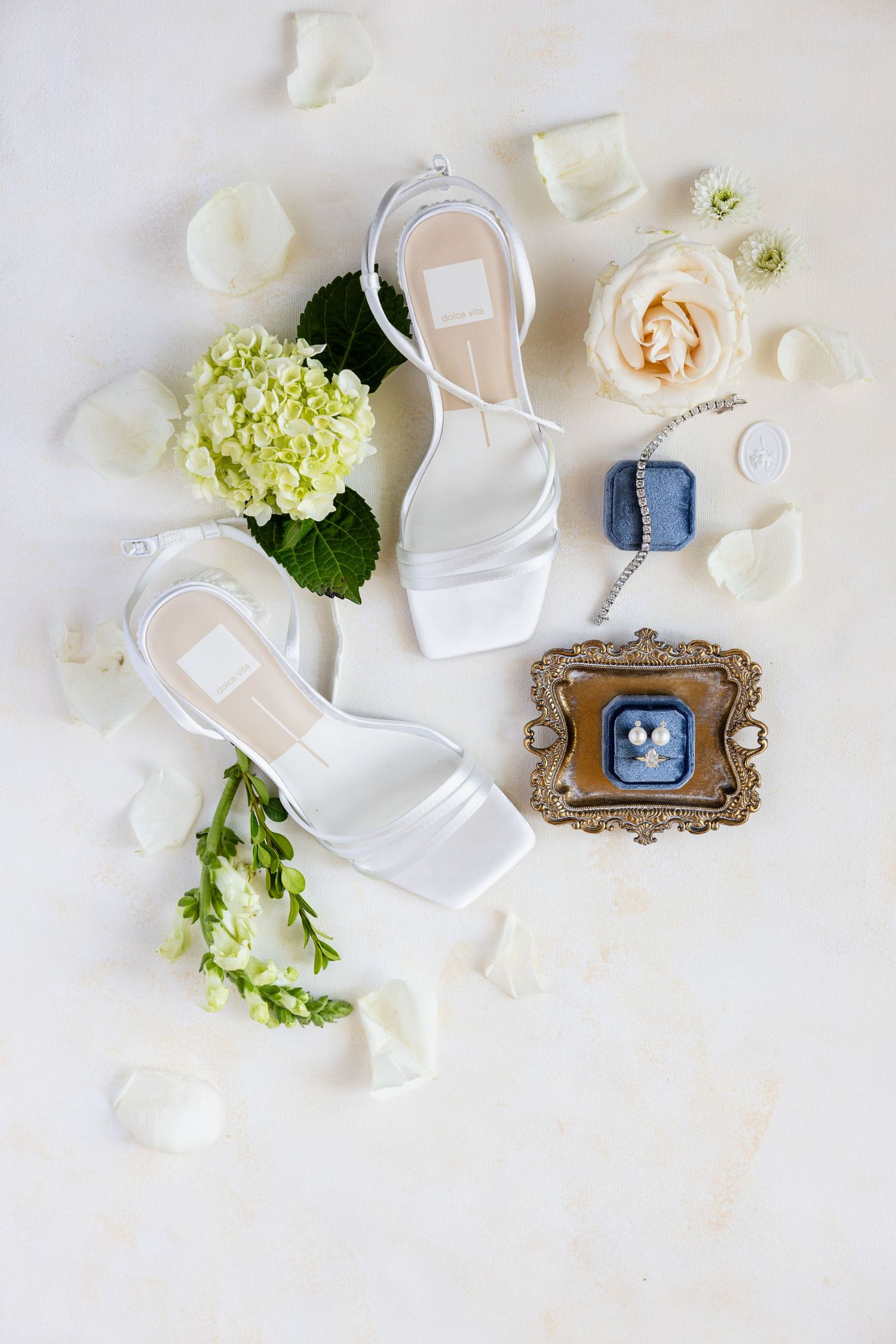 White bridal heels surrounded by hydrangeas, roses, petals, and a blue velvet ring box on a white surface.