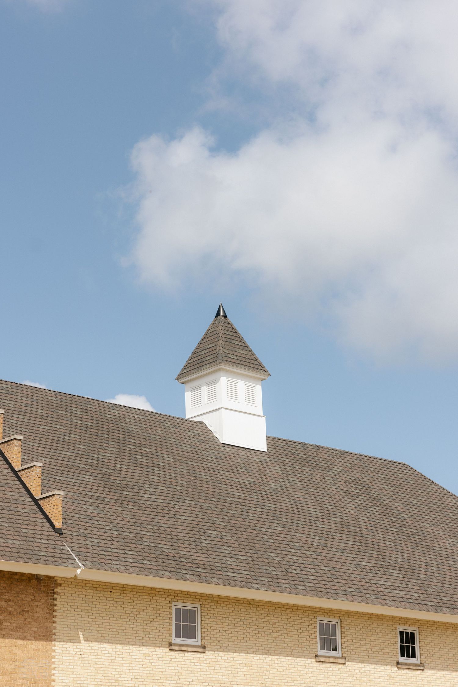 Brick building with white cupola tower on gray shingled roof against blue sky with clouds.