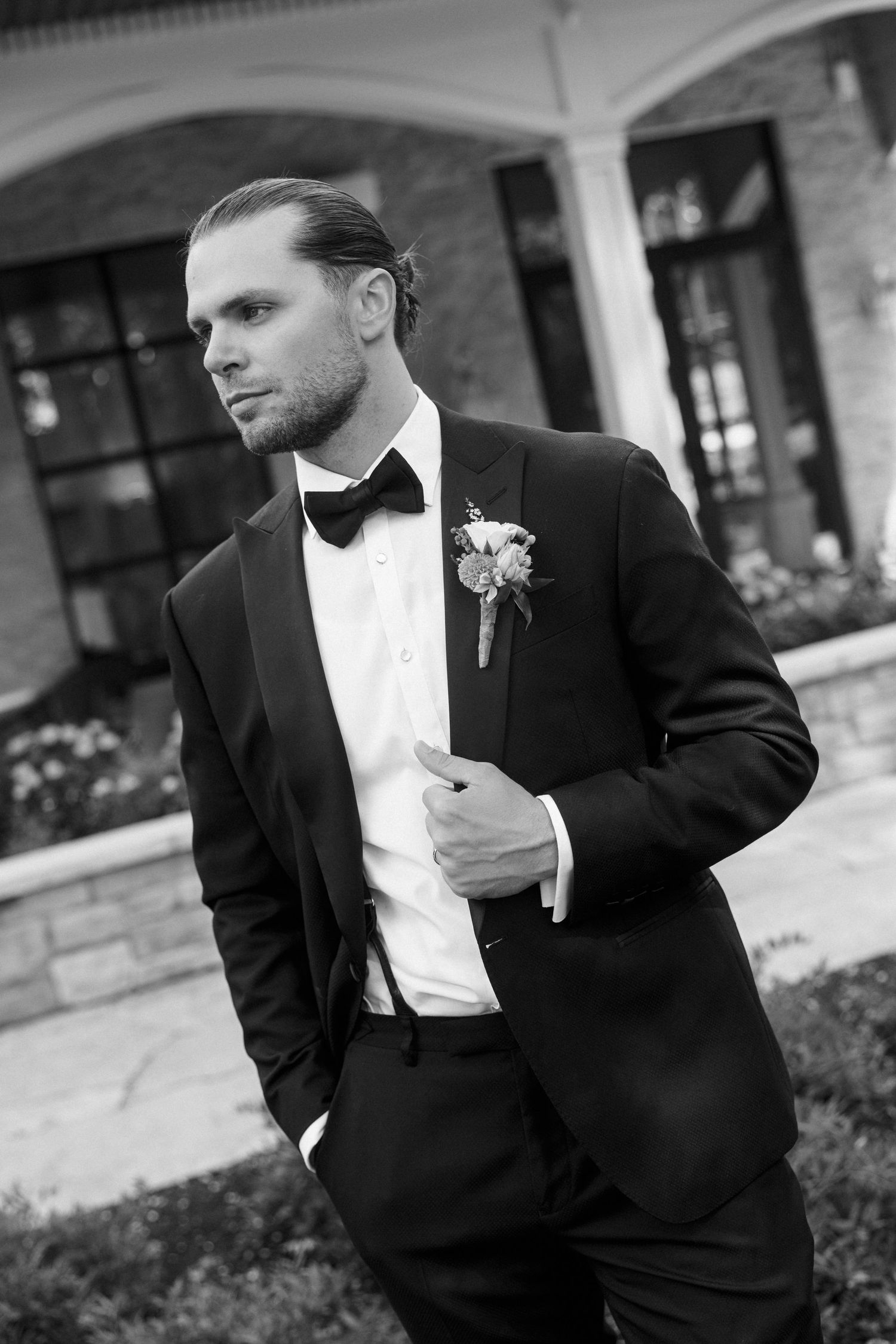 Elegant groom in black tuxedo with bow tie and boutonniere poses outdoors in black and white wedding portrait.