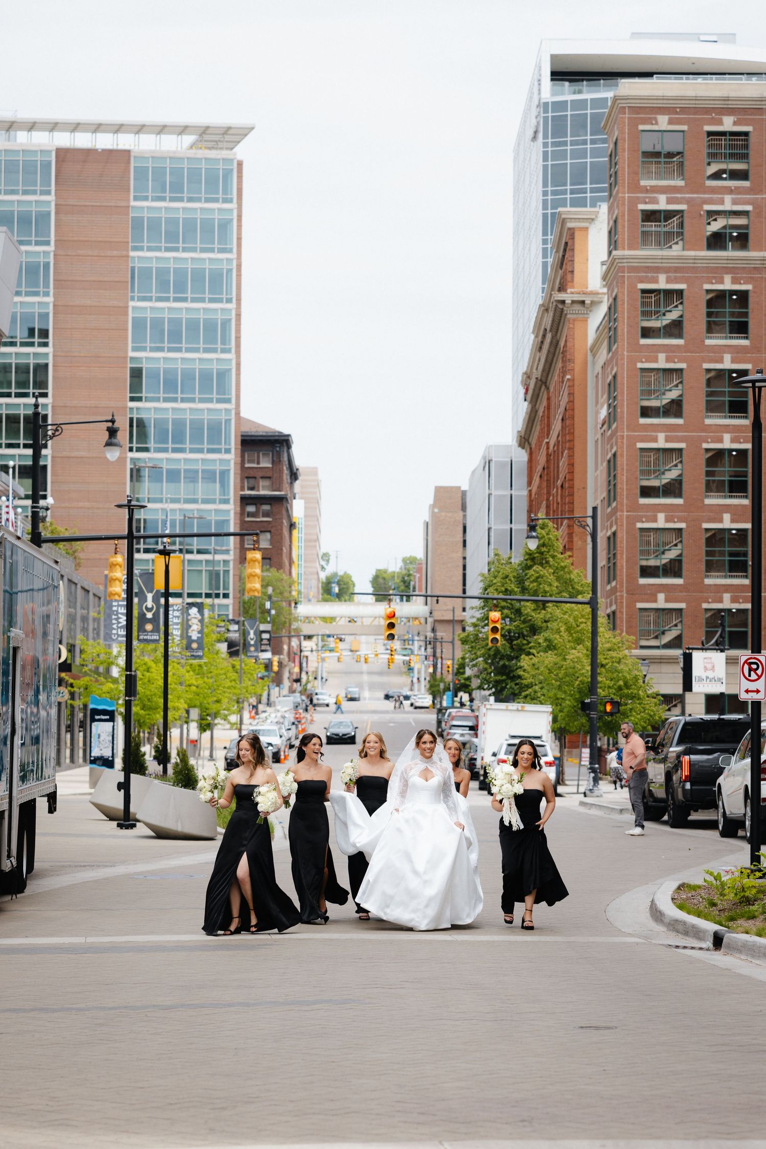 Bride in white gown walks with bridesmaids in black dresses down an urban street lined with modern buildings.