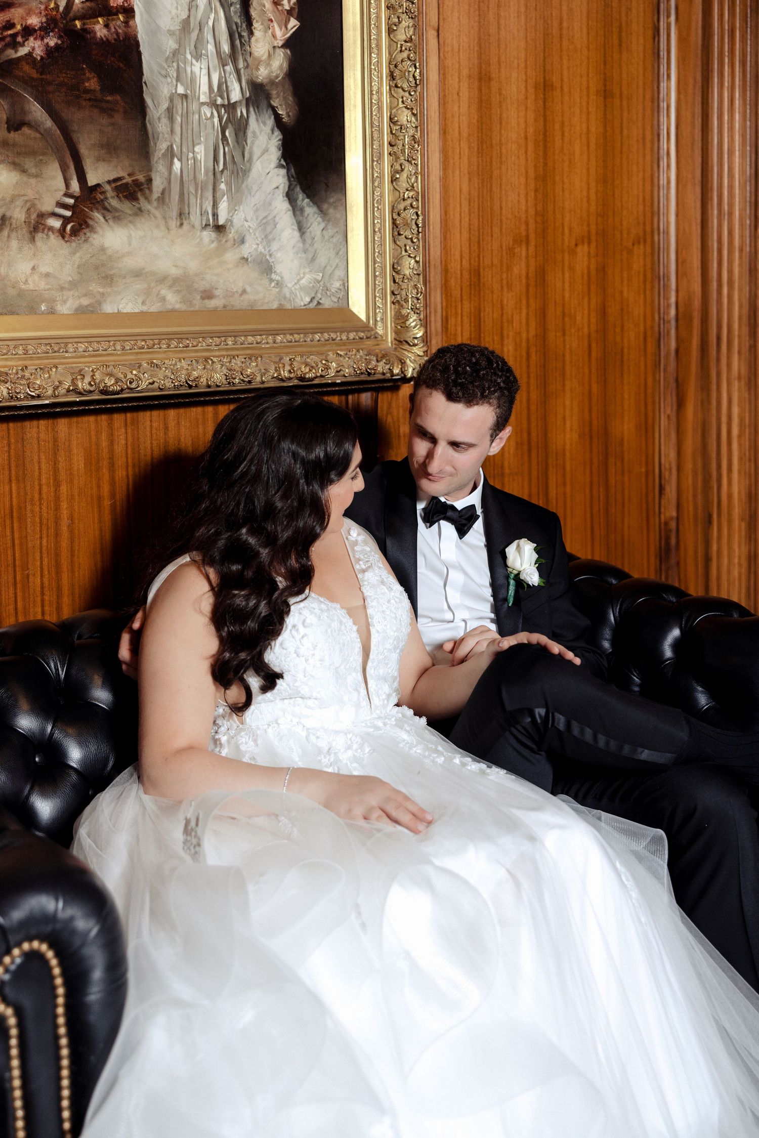 Bride and groom sharing an intimate moment on a tufted leather sofa beneath a gold-framed painting.
