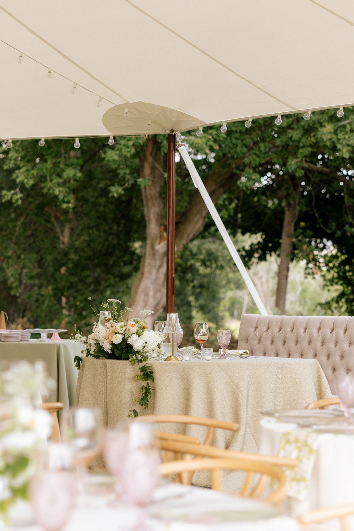 Elegant outdoor wedding reception under a tent with floral centerpieces, tufted seating, and string lights amid lush greenery.