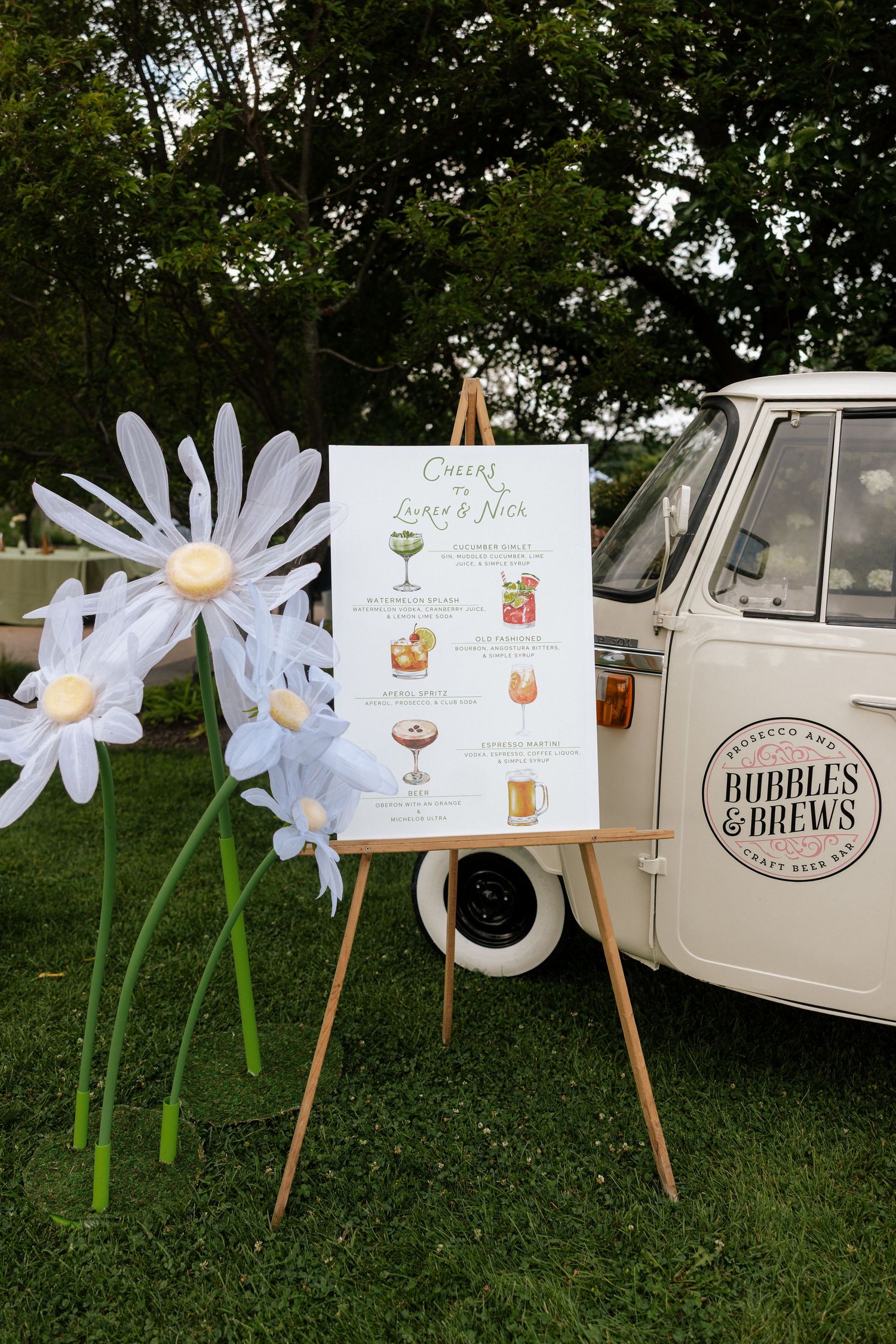 Outdoor wedding cocktail menu on easel beside vintage VW van with giant daisy decorations.