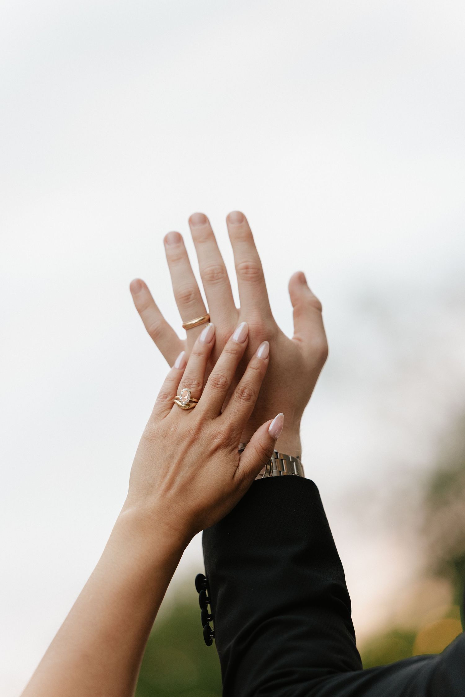 Close-up of two hands raised together, one wearing a gold engagement ring and the other a silver watch, against a cloudy sky.