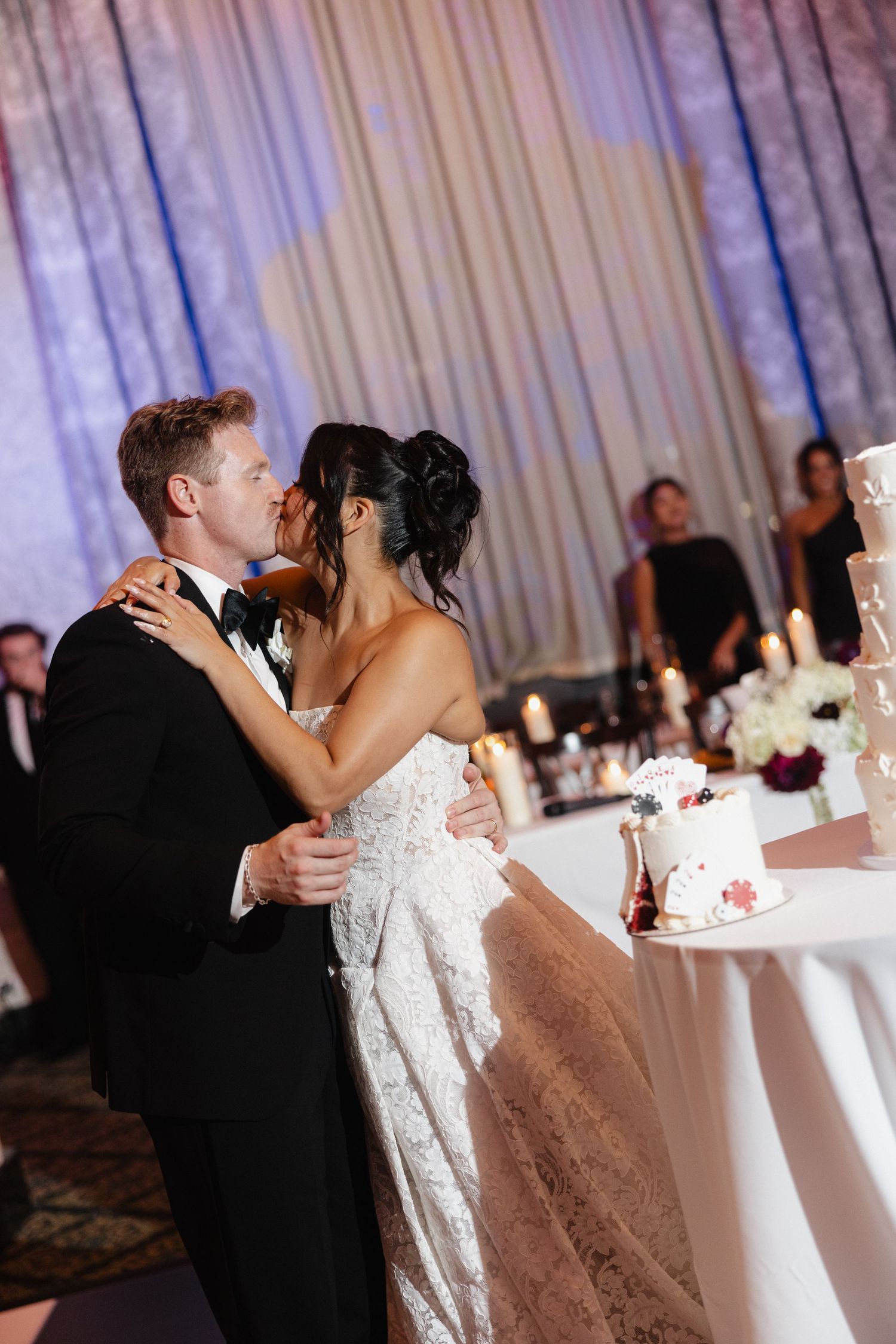 Bride and groom share a romantic kiss at their wedding reception near a decorated cake table with candles.
