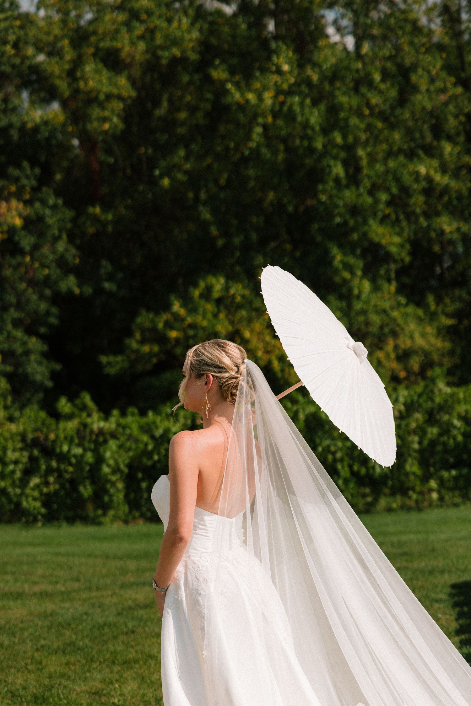 Bride in white gown holding a white parasol with flowing veil in a lush green garden setting.