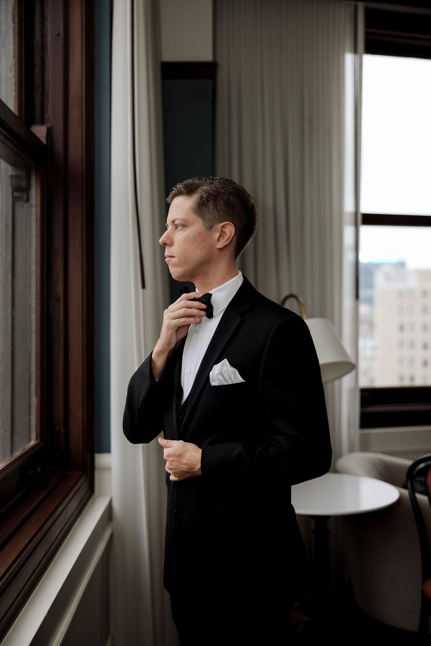 A groom in a black tuxedo adjusts his bow tie while gazing out a hotel window before his wedding ceremony.