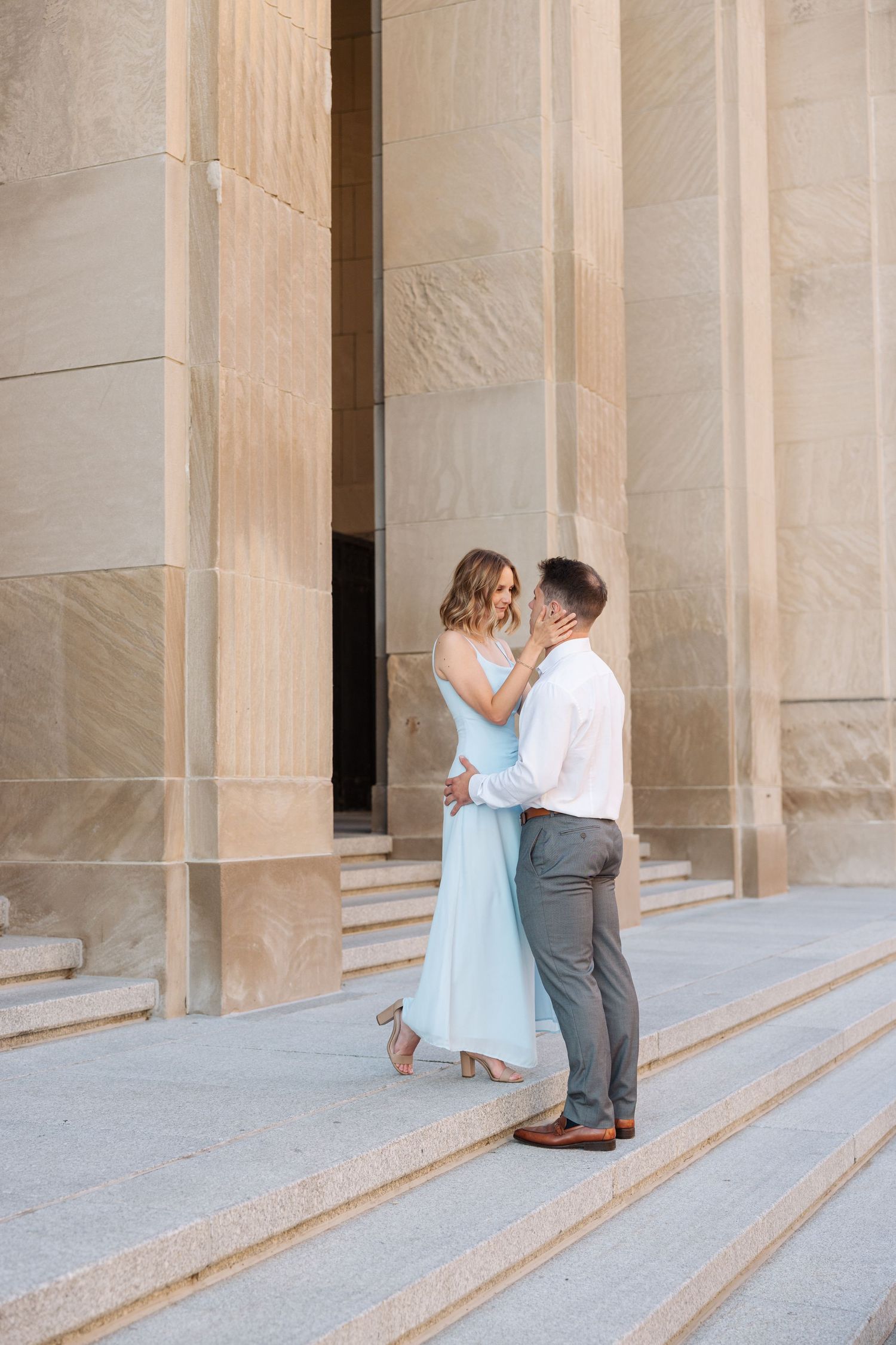 Couple in elegant attire embracing on stone steps before grand columns in a romantic outdoor setting.