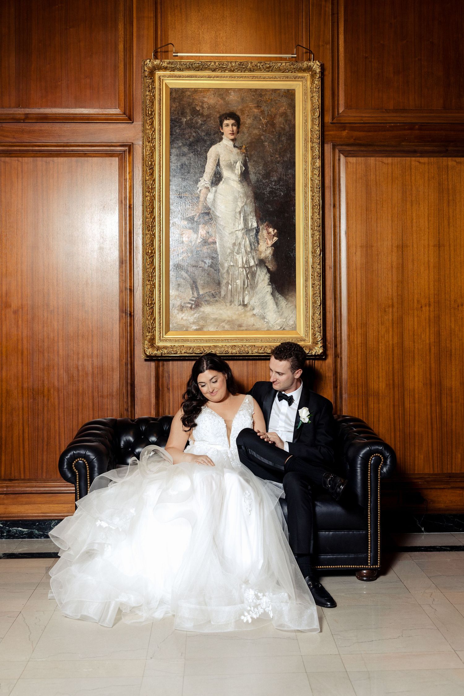 Bride and groom sitting on a black leather sofa beneath a classic portrait painting in an elegant wood-paneled room.