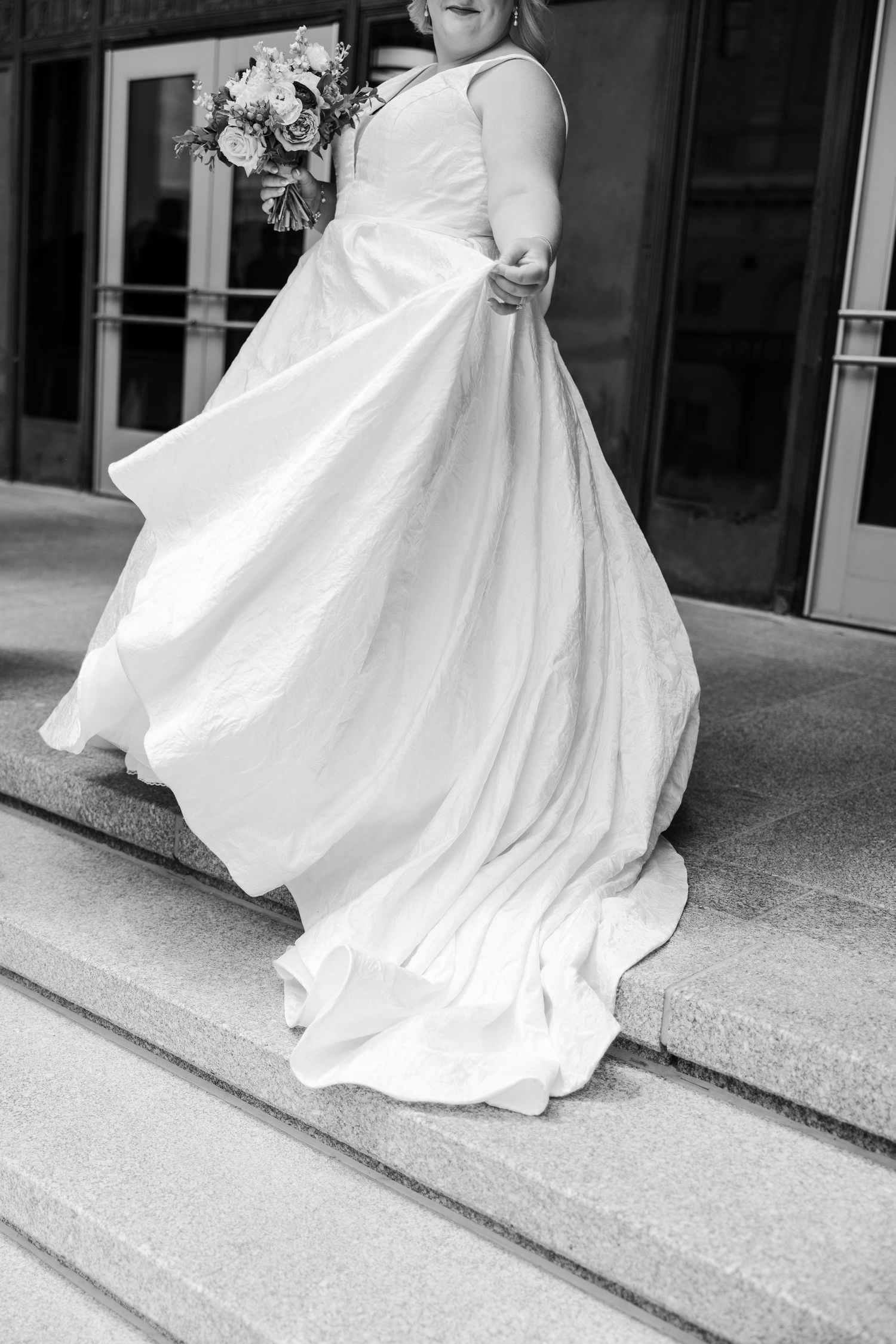 Black and white photo of a bride in a flowing white wedding gown holding a bouquet while descending outdoor steps.