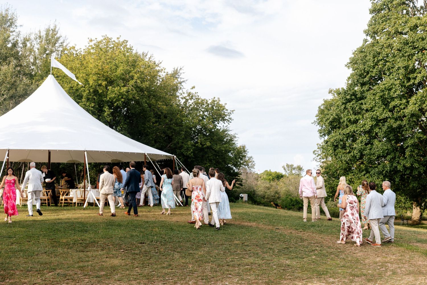 Guests in elegant attire gather on a green lawn near a white tent at an outdoor wedding reception.