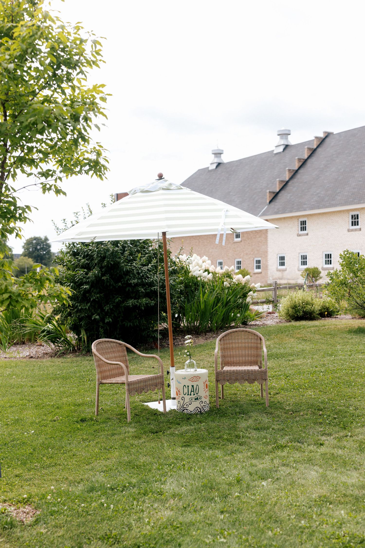 Two wicker chairs and striped patio umbrella on green lawn with garden and barn building in background.