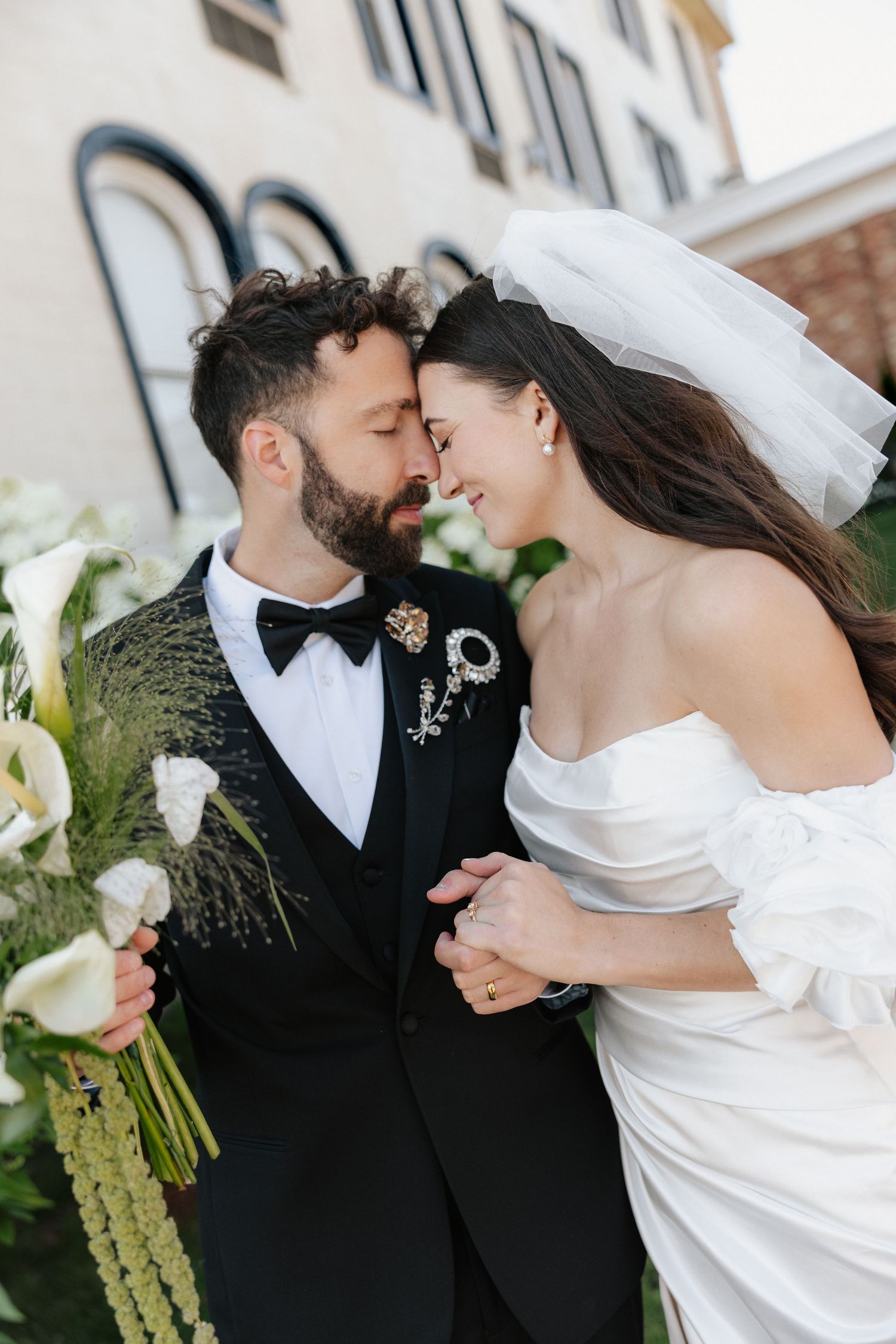 Bride and groom share a romantic moment, she in a white off-shoulder gown with veil, he in a black tuxedo with bow tie.