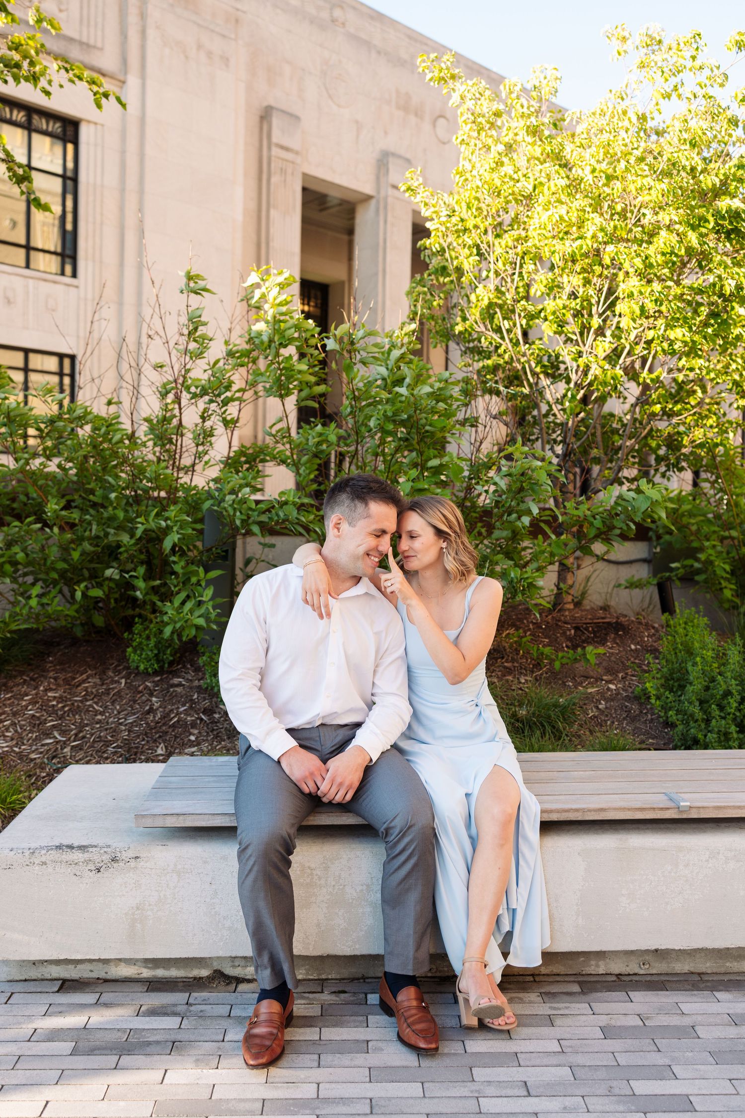Couple sitting close together on a stone bench in a sunlit garden outside a classic building.