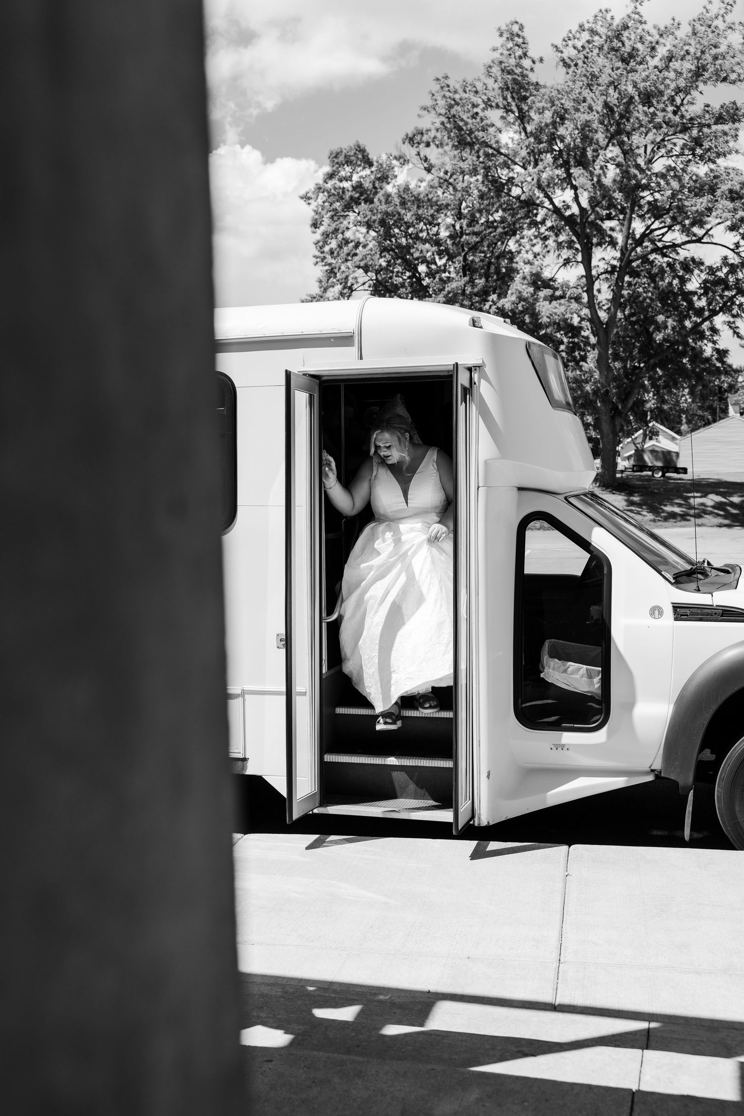Bride in white wedding dress stepping out of a white shuttle bus on her wedding day, black and white photo.