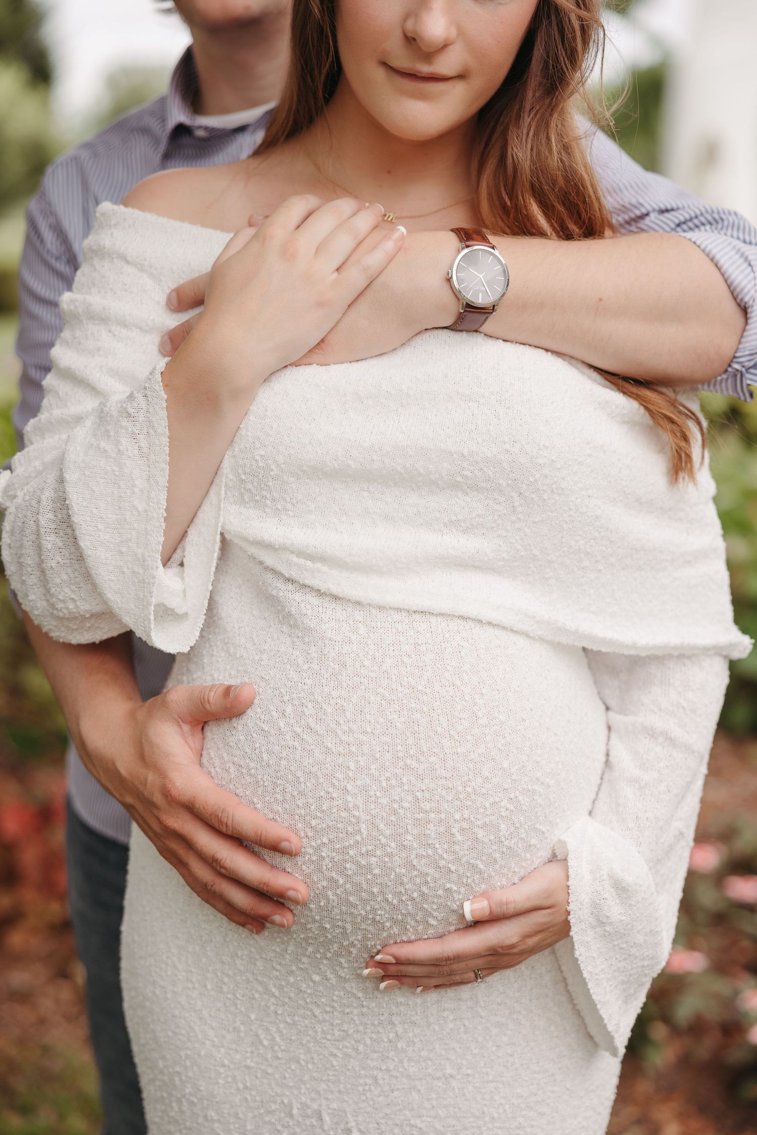 Pregnant woman in white off-shoulder dress cradled by partner outdoors.