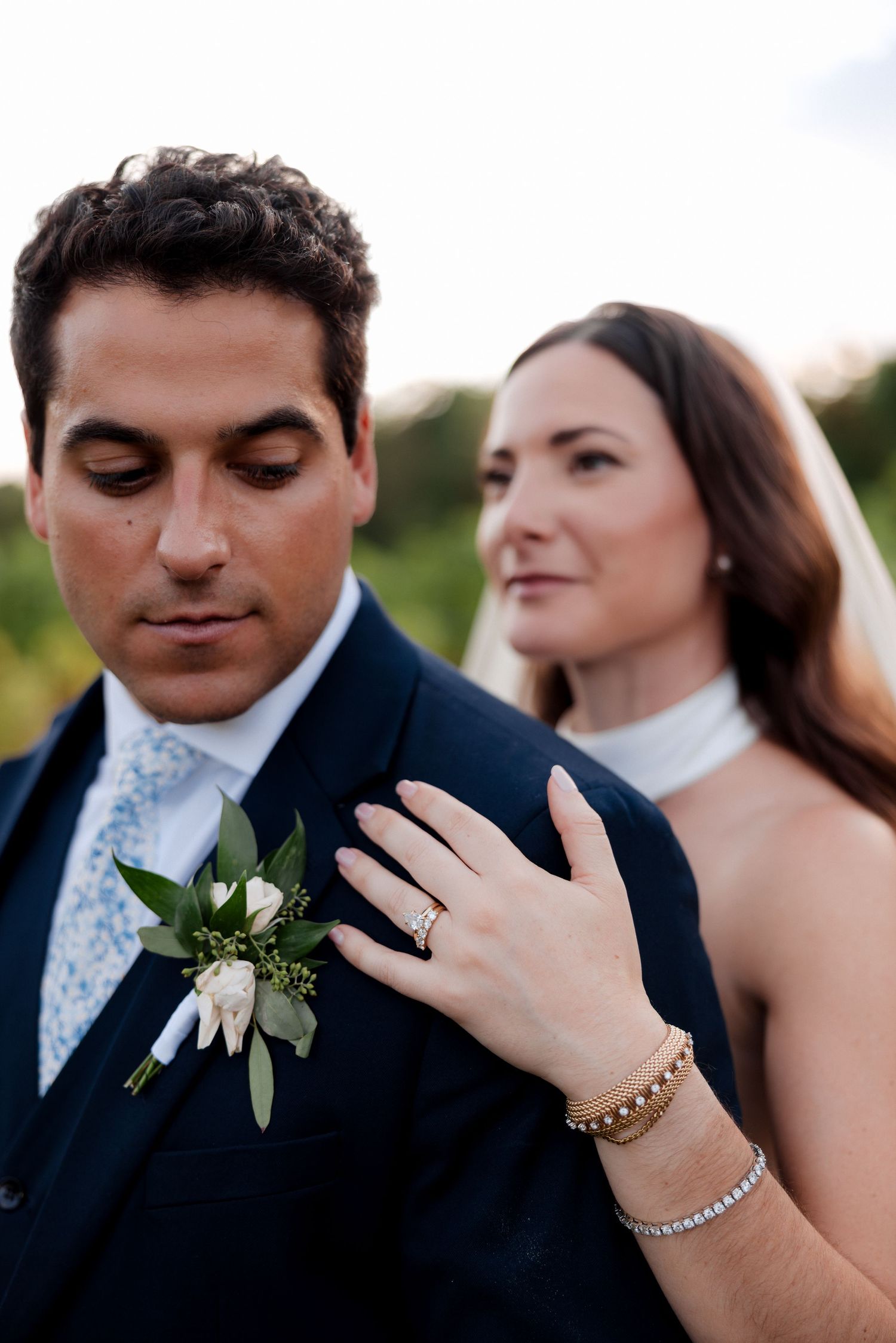 Bride places hand on groom's shoulder showing diamond ring, groom wears navy suit with white boutonniere at outdoor wedding.
