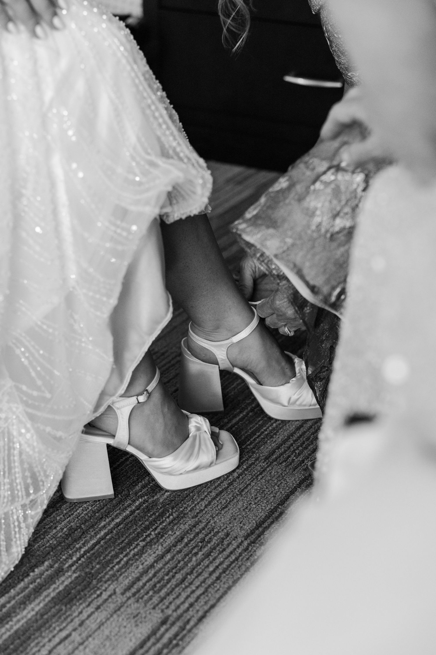 Black and white photo of a bride wearing elegant white block heel sandals with bow detail on a carpeted floor.
