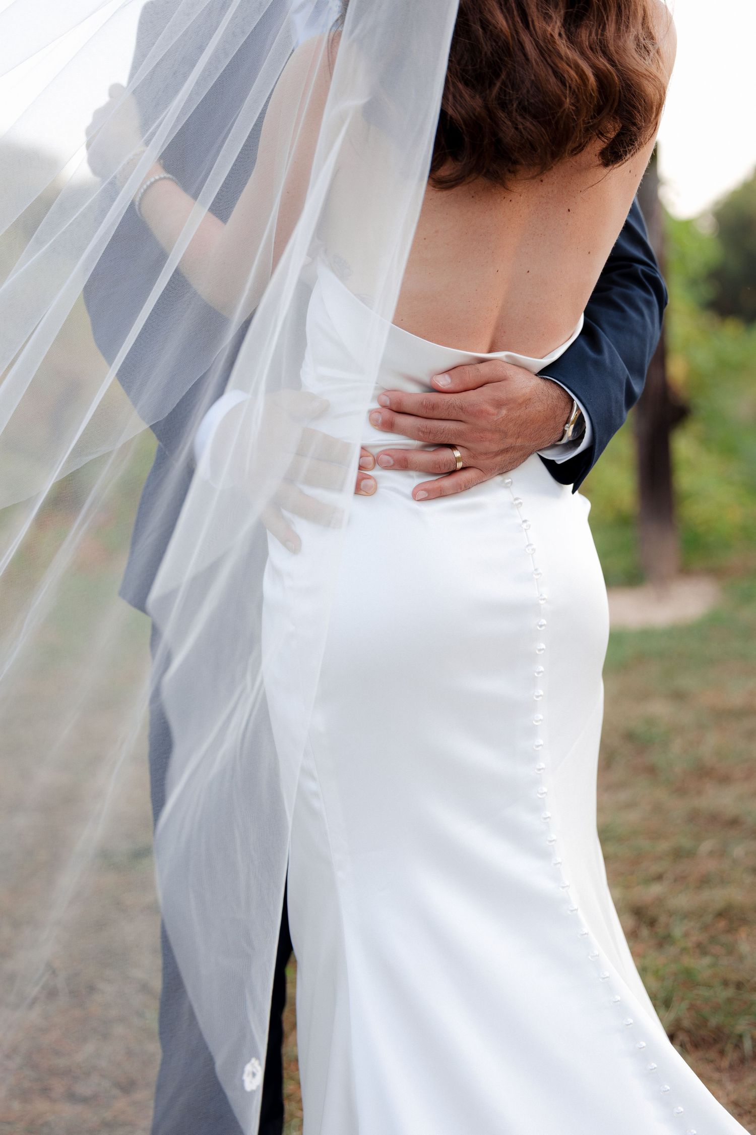 Groom embracing bride from behind, showcasing backless white satin gown with veil and button details.