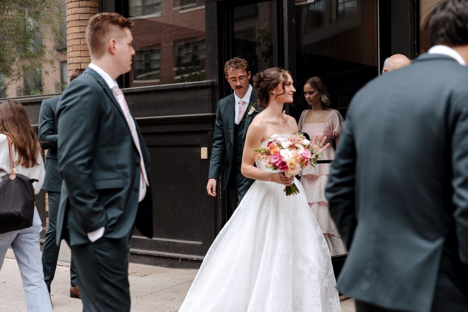 Smiling bride holding colorful bouquet mingles with wedding guests outside dark storefront.