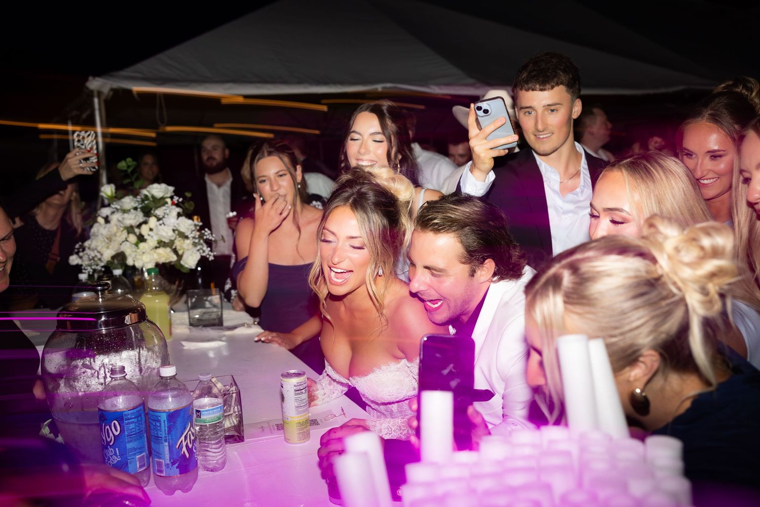Bride and groom joyfully celebrating with guests at a lively wedding reception under purple lights.