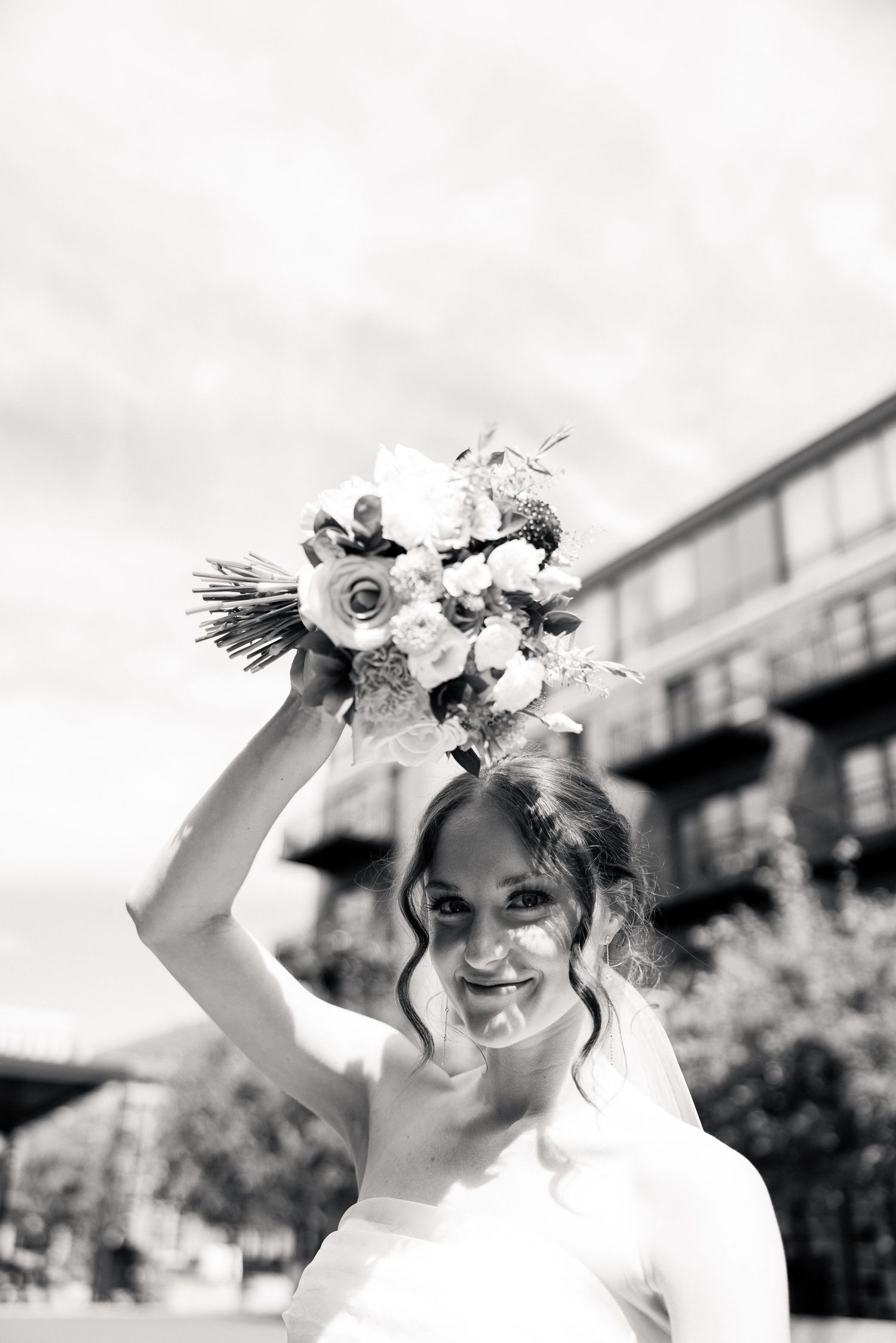 Smiling bride holding floral bouquet raised overhead in black and white portrait outside urban wedding venue.