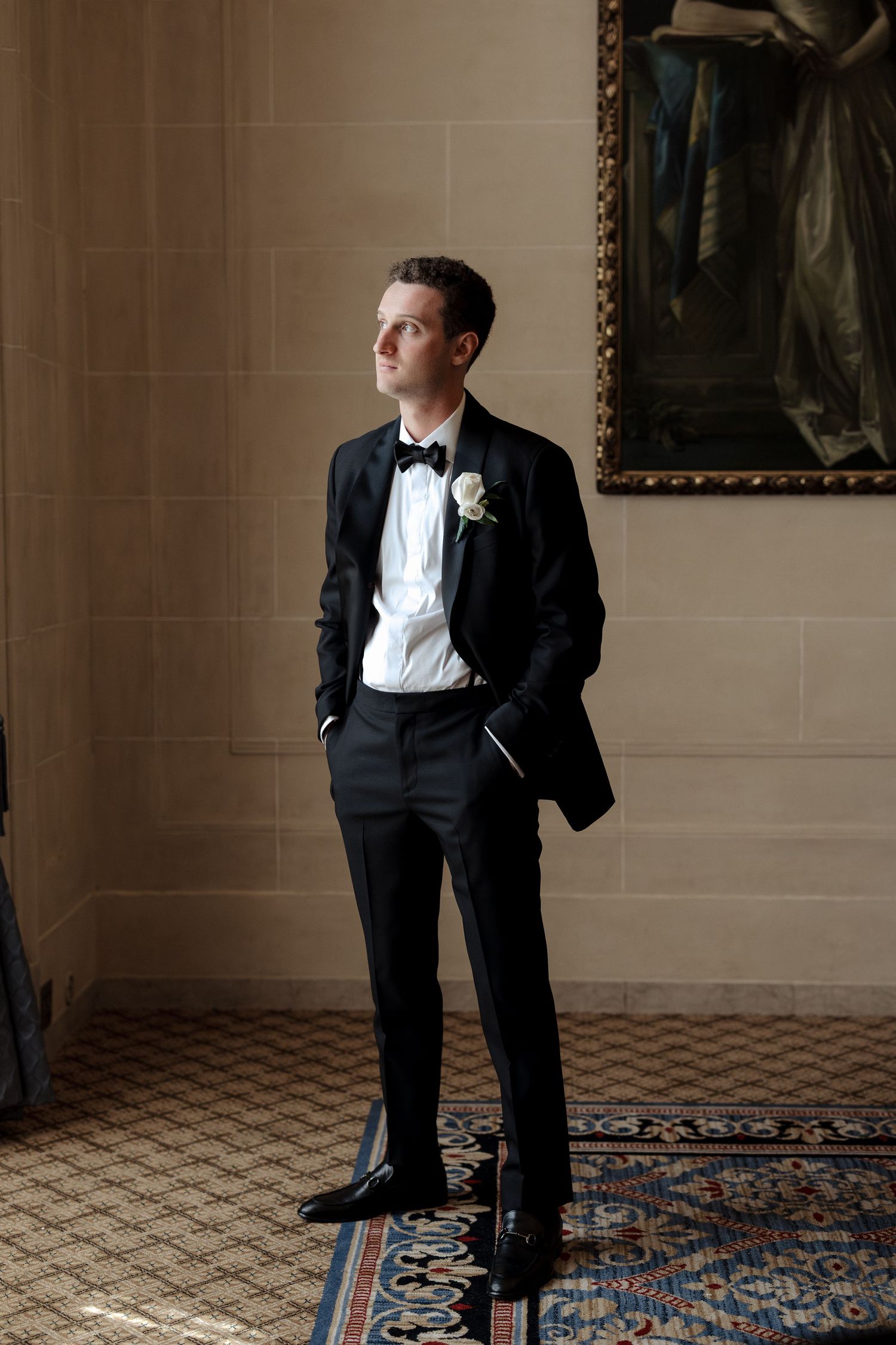 Groom in black tuxedo with white boutonniere standing confidently in an elegant hall with ornate painting.