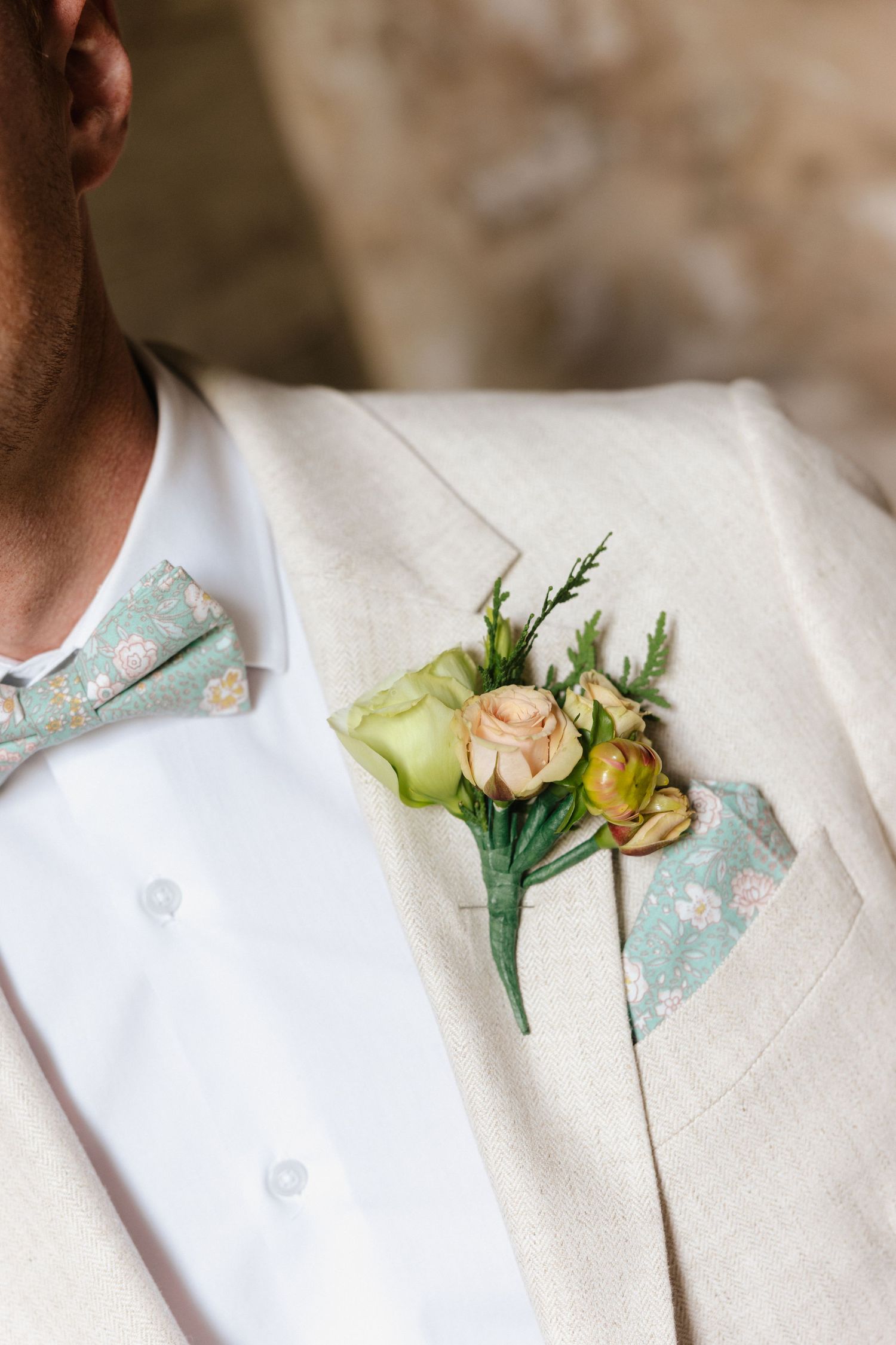 Groom wearing beige linen suit with floral boutonniere and mint floral bow tie at wedding.