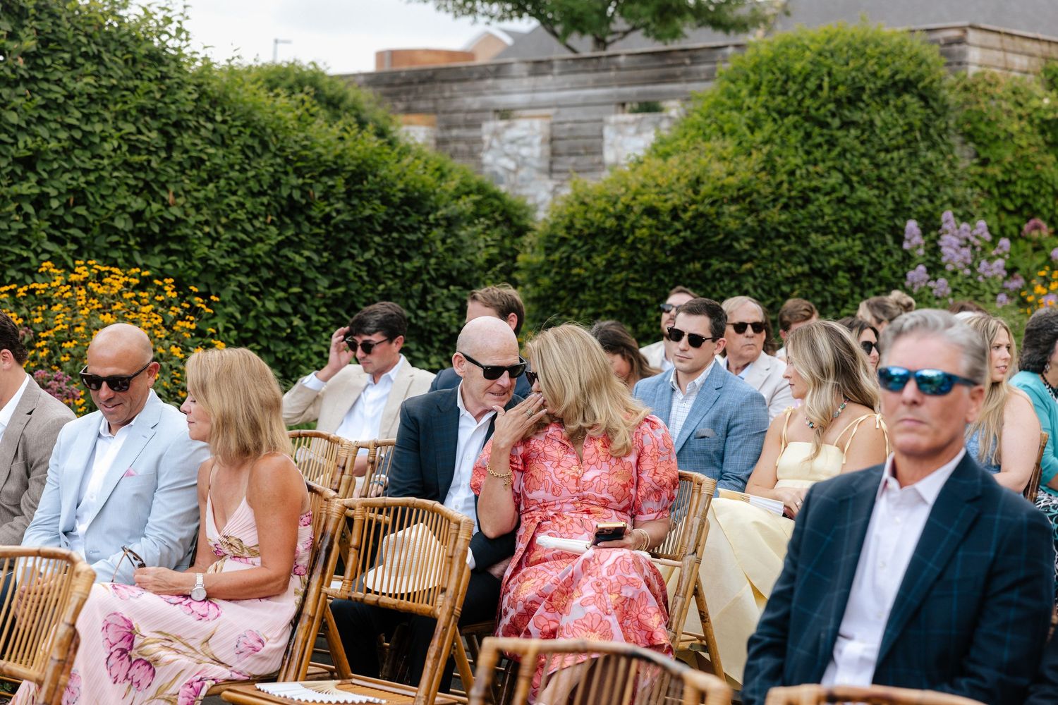 Guests in sunglasses seated in rattan chairs at an outdoor wedding ceremony surrounded by lush green hedges.