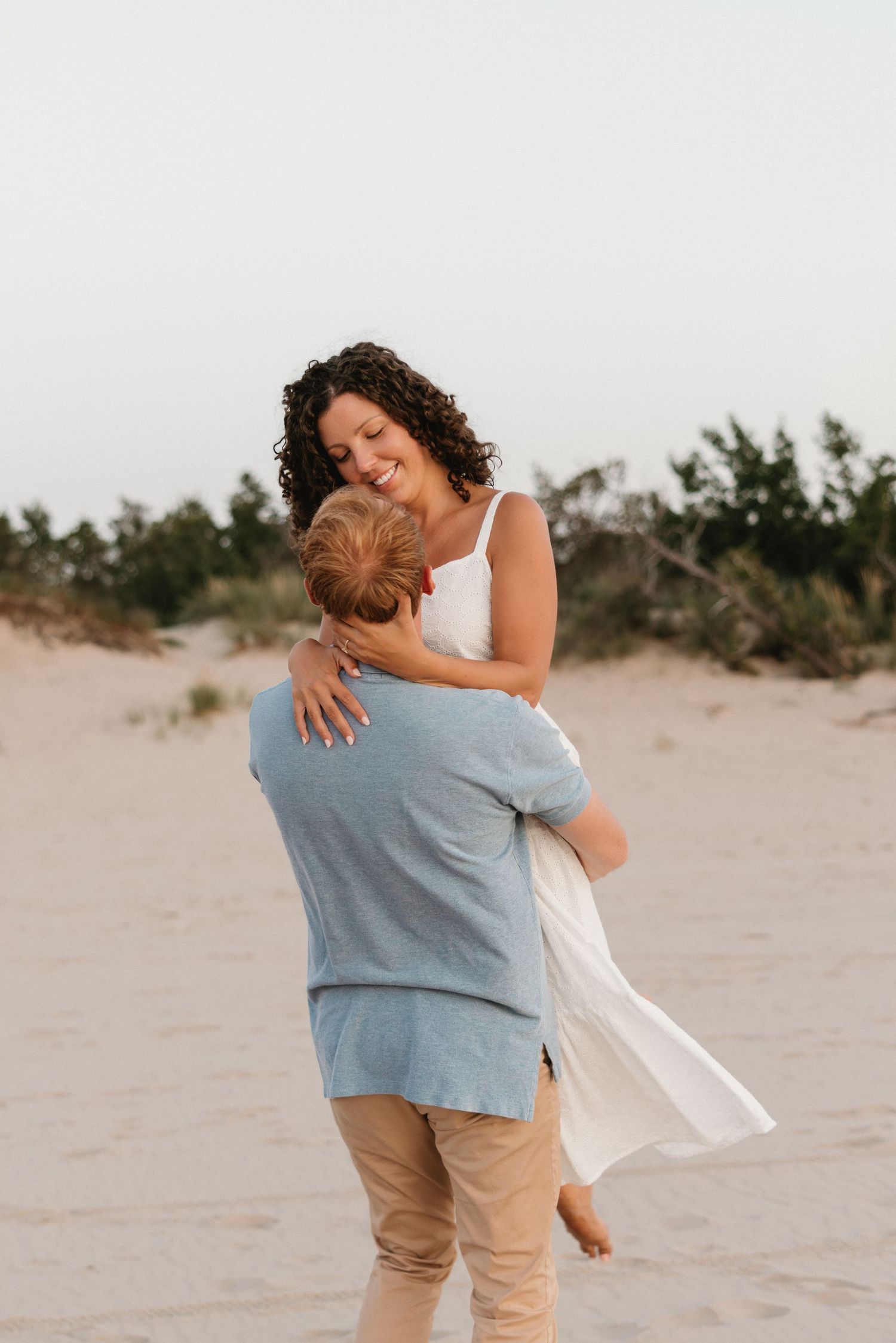A couple embraces on a sandy beach, the woman in a white dress smiling joyfully as the man lifts her up.
