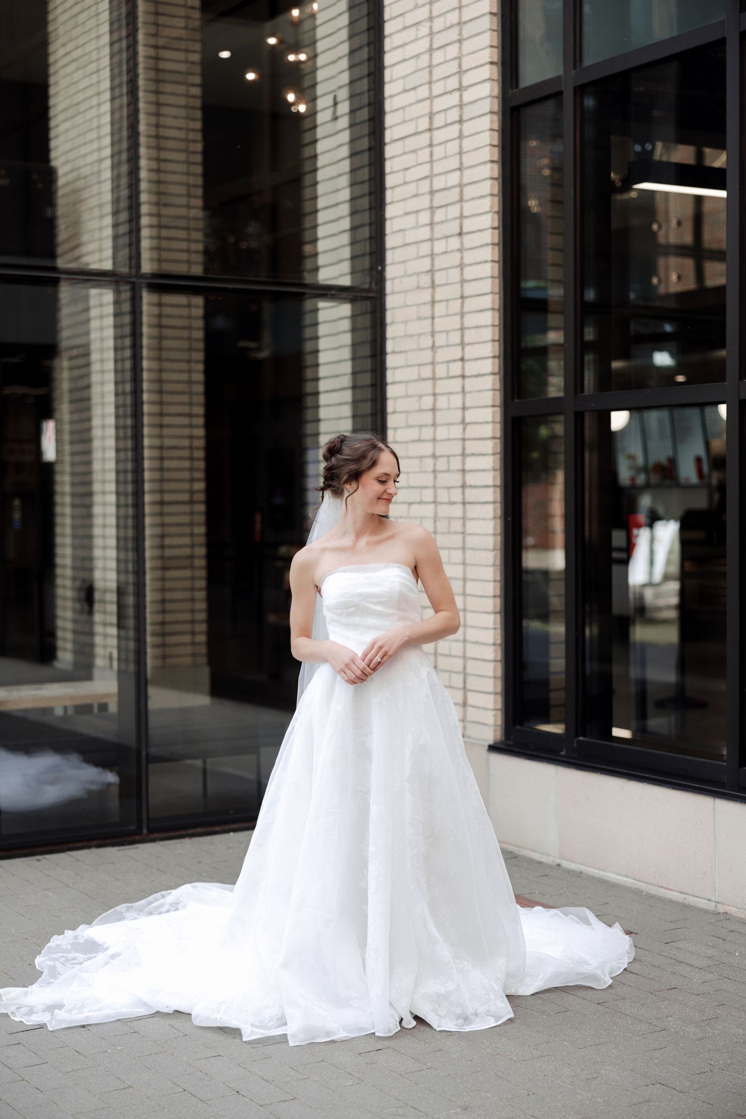 Bride in strapless white ballgown with veil stands outside modern brick building on wedding day.