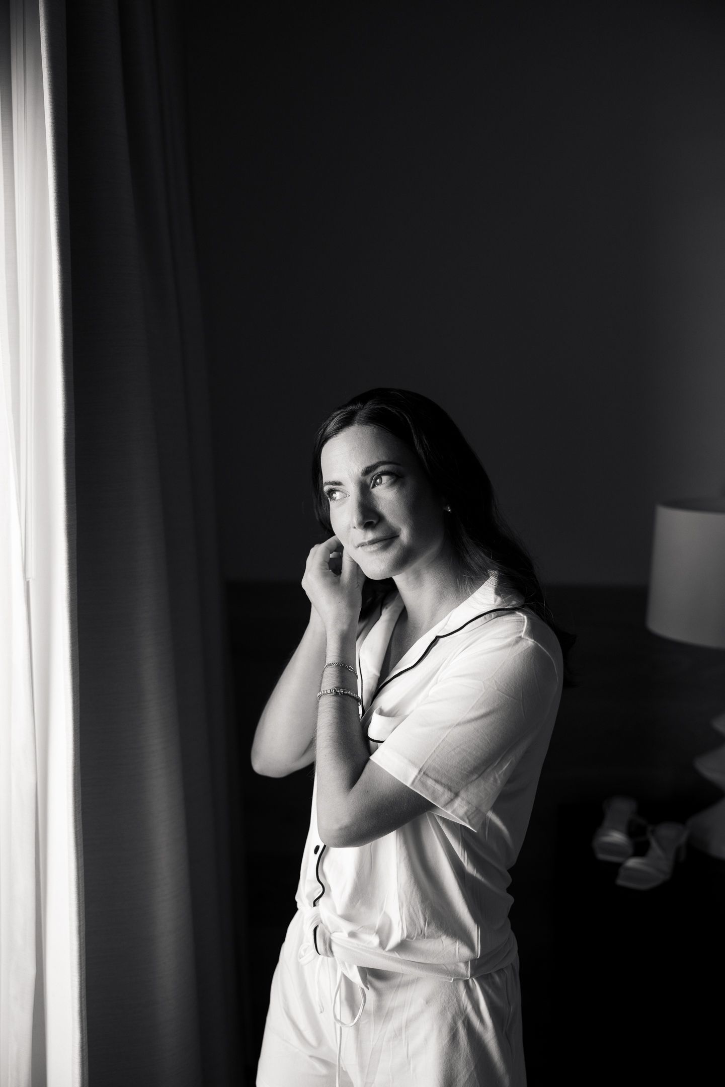 Black and white portrait of a smiling woman in white pajamas standing by a window in a dimly lit bedroom.