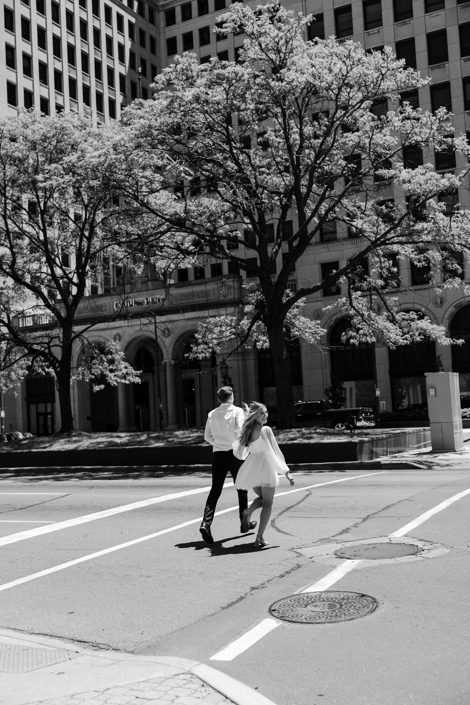 Black and white photo of a couple crossing a sunny city street with flowering trees in bloom.