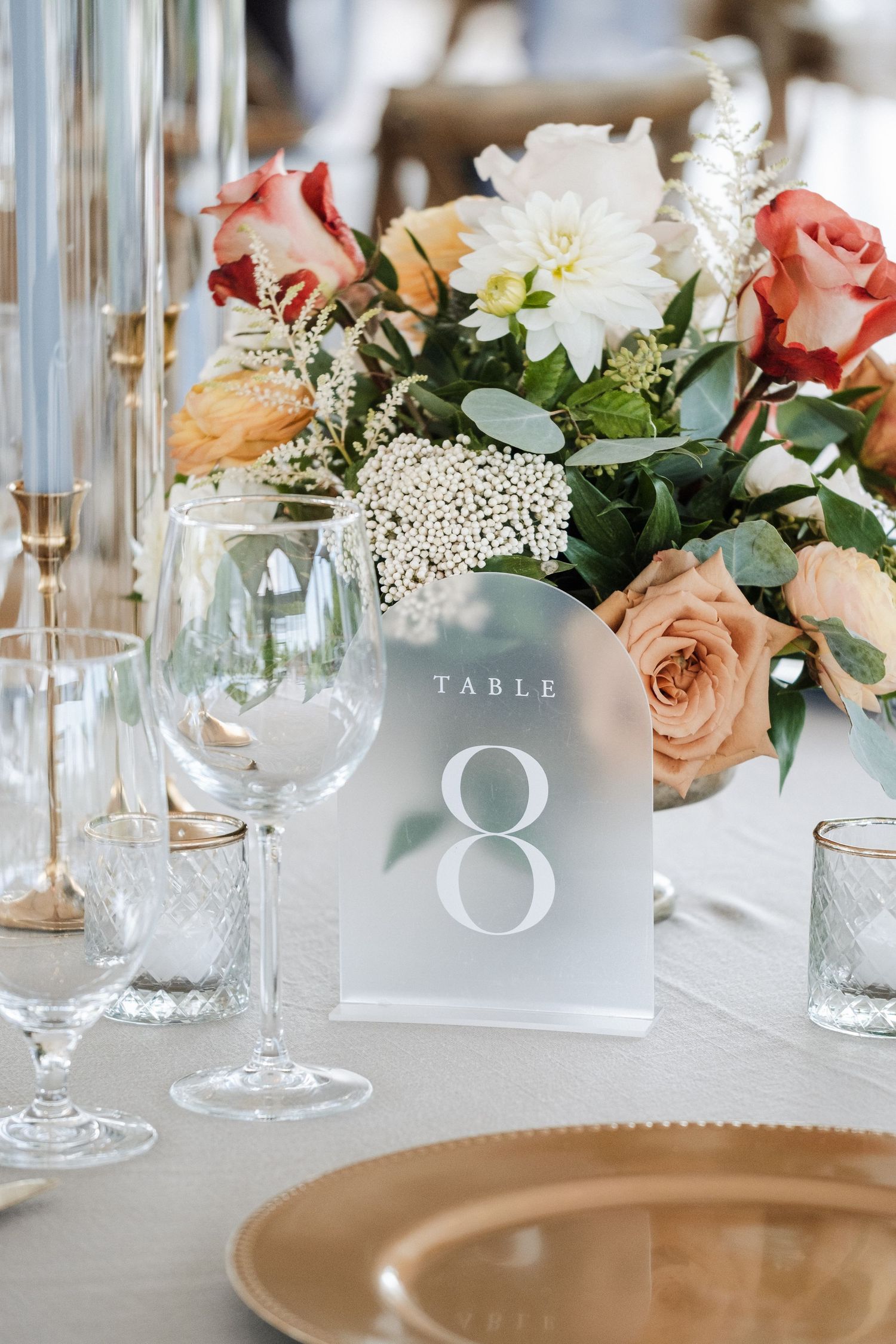 Elegant wedding table setting with Table 8 sign, peach and red floral centerpiece, crystal glassware, and gold charger plates.