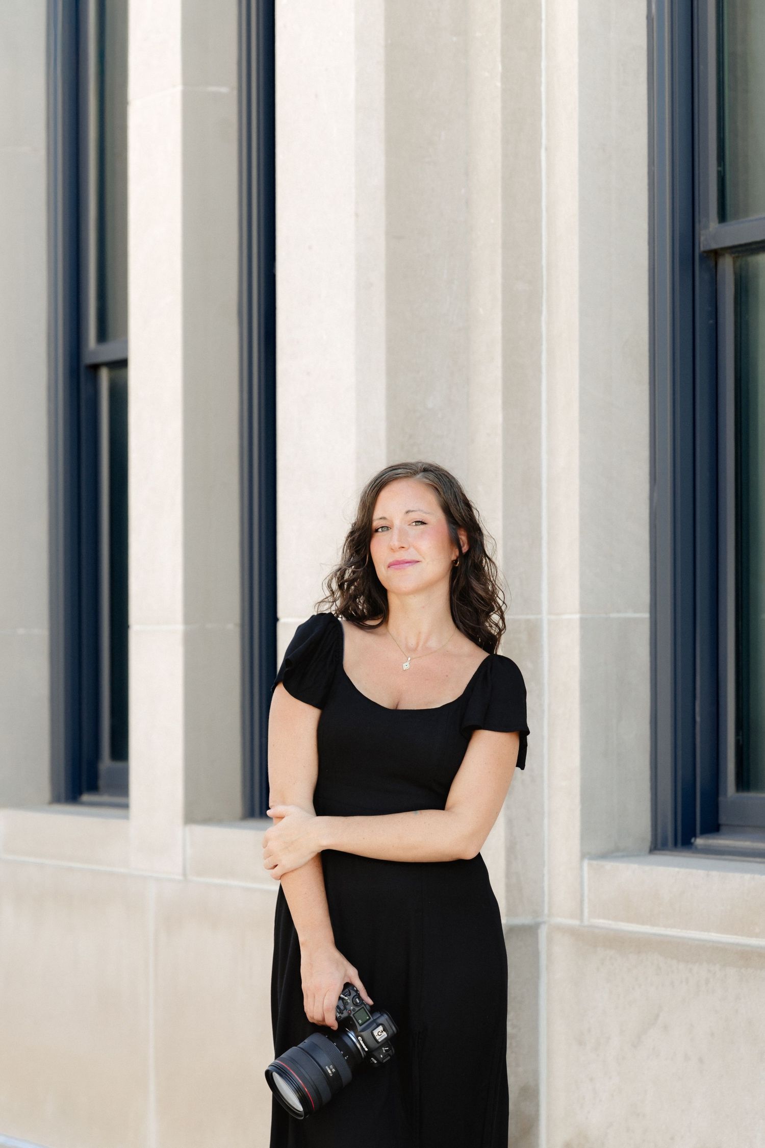 A woman in a black dress holds a DSLR camera while posing confidently beside stone columns outdoors.