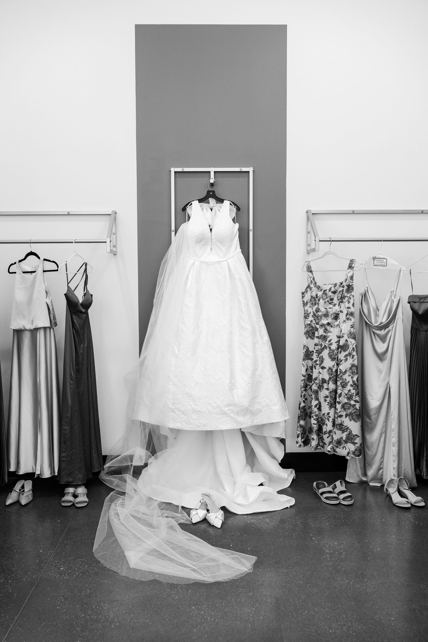 Black and white photo of a wedding dress on display flanked by bridesmaid dresses on racks with shoes below.
