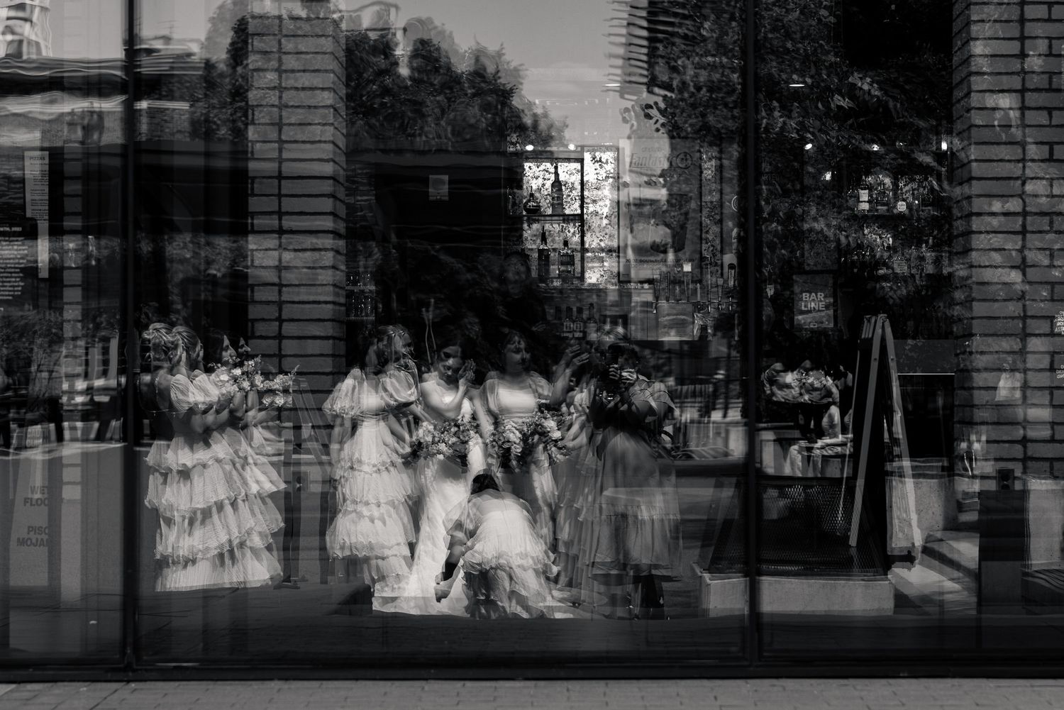 Artistic black and white reflection of bridesmaids in ruffled dresses captured through large glass window pane.