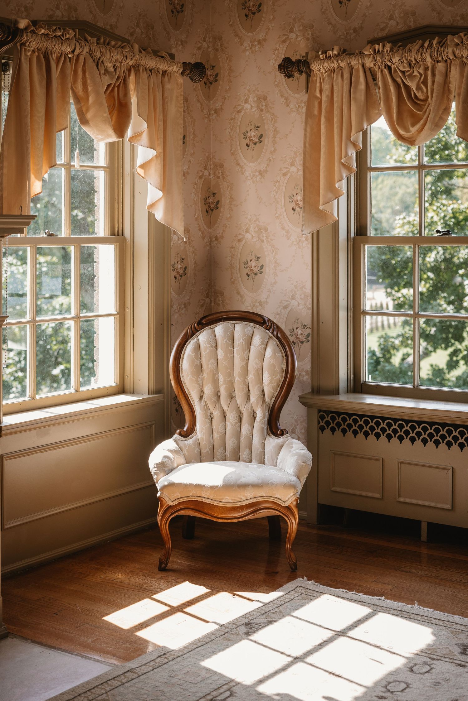 Antique white tufted armchair bathed in natural light beside two large windows with gold curtains.