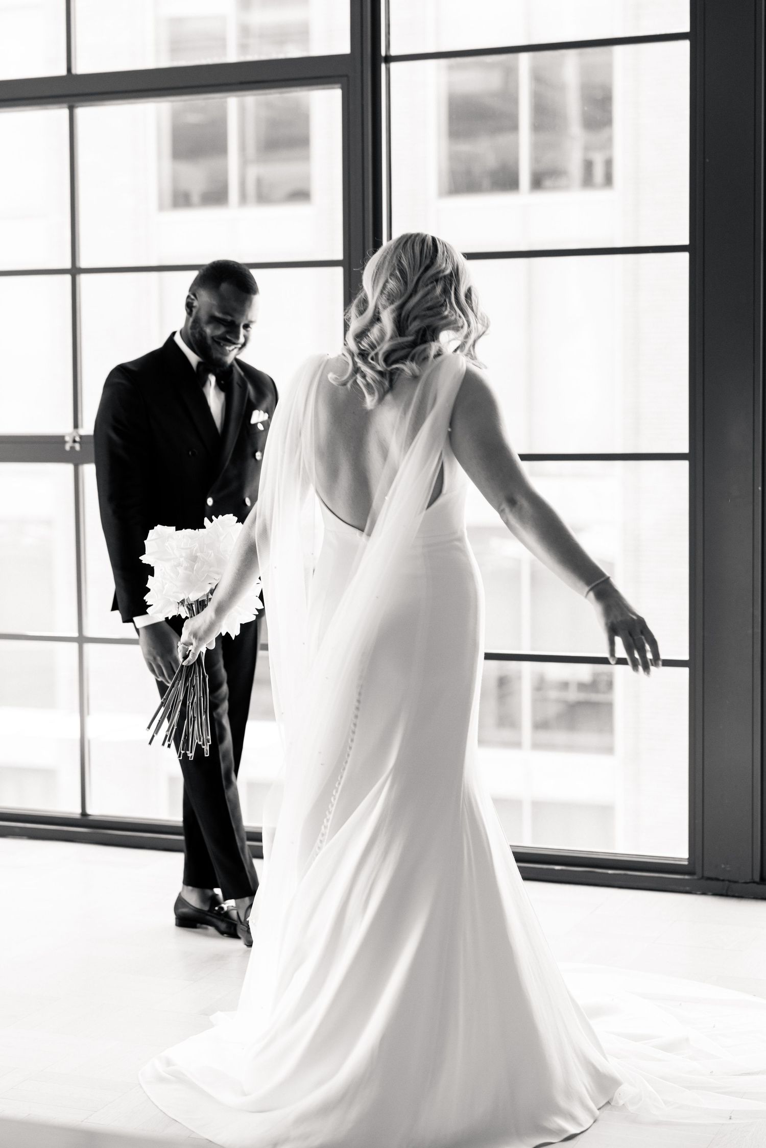 Black and white first look photo of groom smiling at bride in elegant backless gown by large windows.
