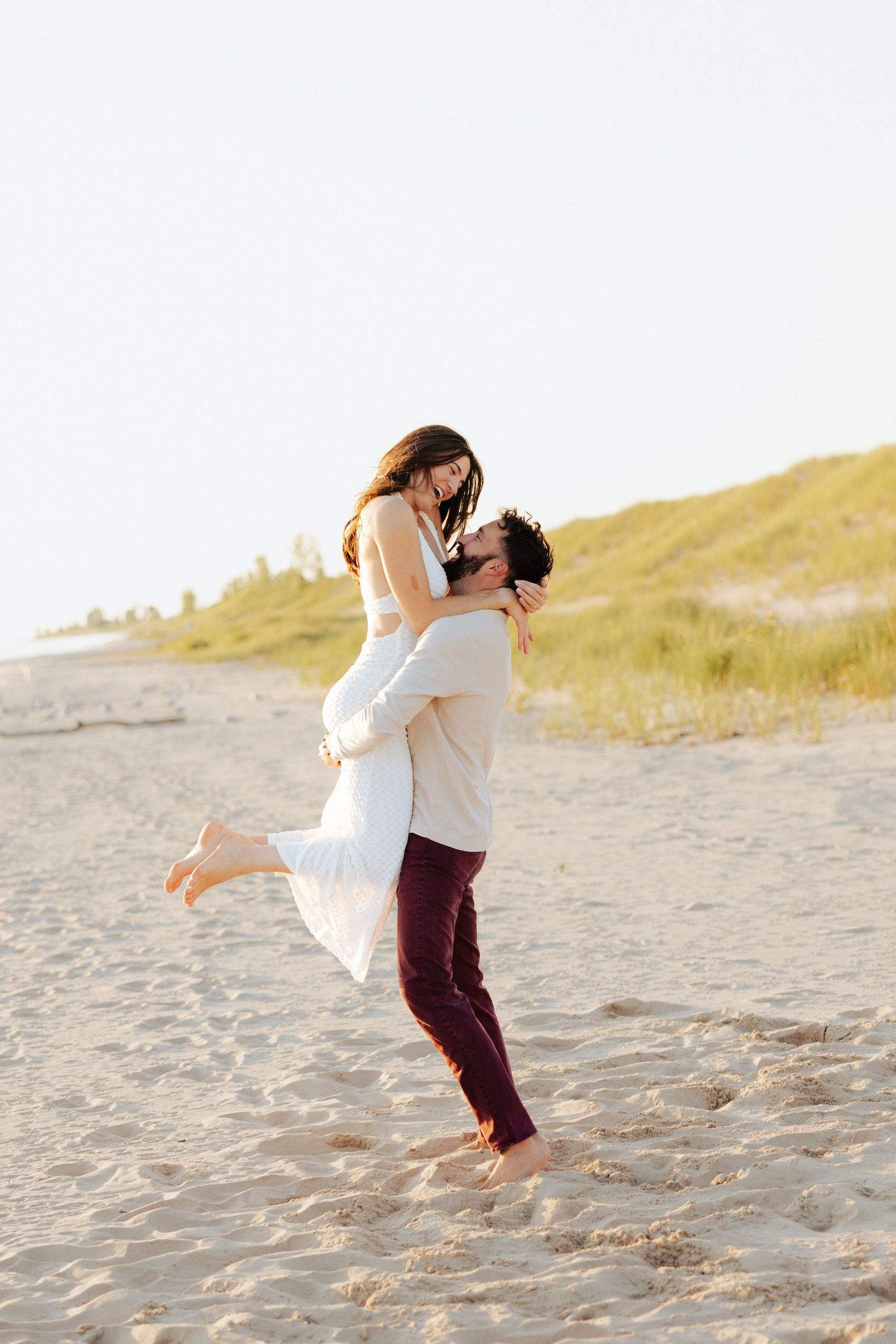 A couple shares a romantic moment on a sandy beach, as the man lifts the woman in a white dress at sunset.