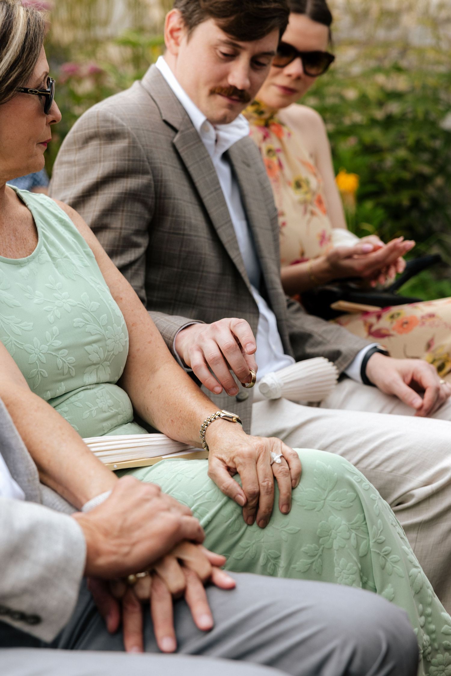Wedding guests seated outdoors in garden ceremony, holding fans and programs during service.