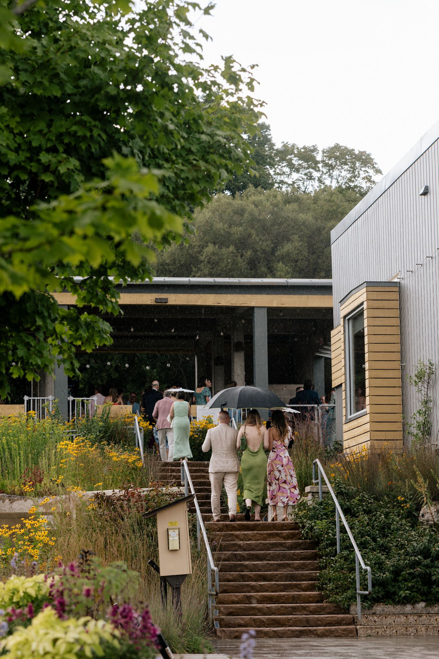 Guests walk up stone steps toward a rustic venue surrounded by lush gardens, some carrying umbrellas on a cloudy day.