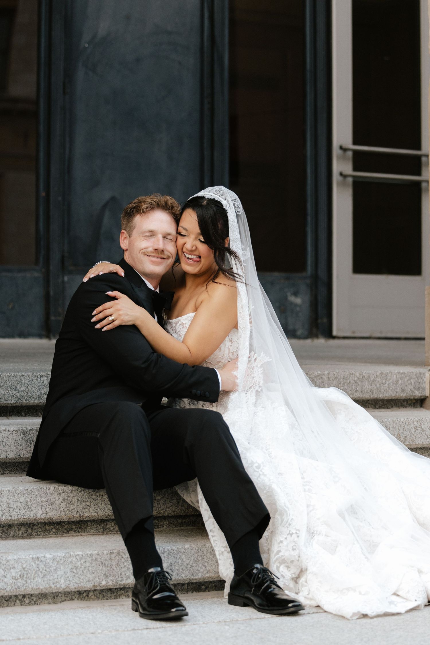 A bride in a lace gown and veil hugs a groom in a black tuxedo as they sit together on stone steps.