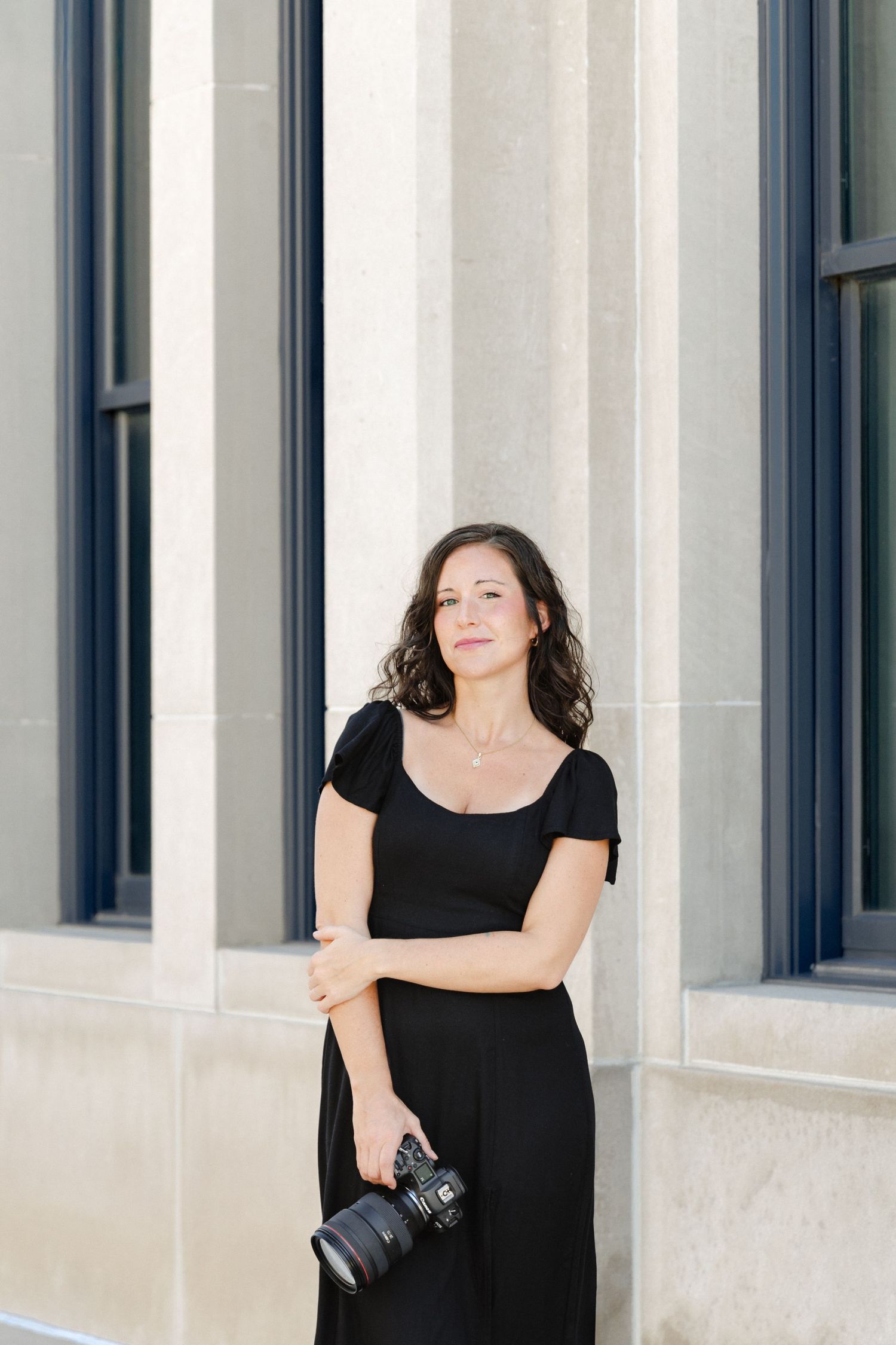 A woman in a black dress holds a DSLR camera while posing elegantly beside stone columns outdoors.